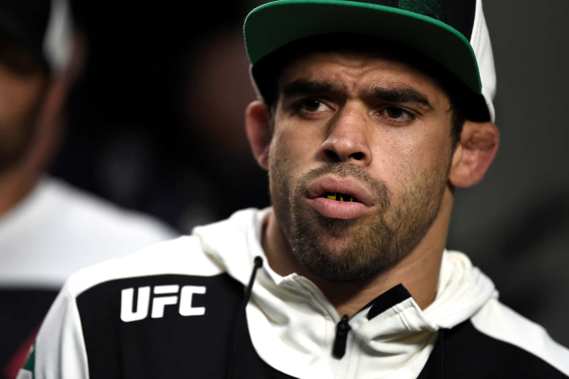 巴西綜合格鬥選手Renan Barão戴帽穿UFC外套近照 壁紙 — 專注的UFC拳手特寫，戴著綠色和白色帽子及品牌連帽衫，背景深色。