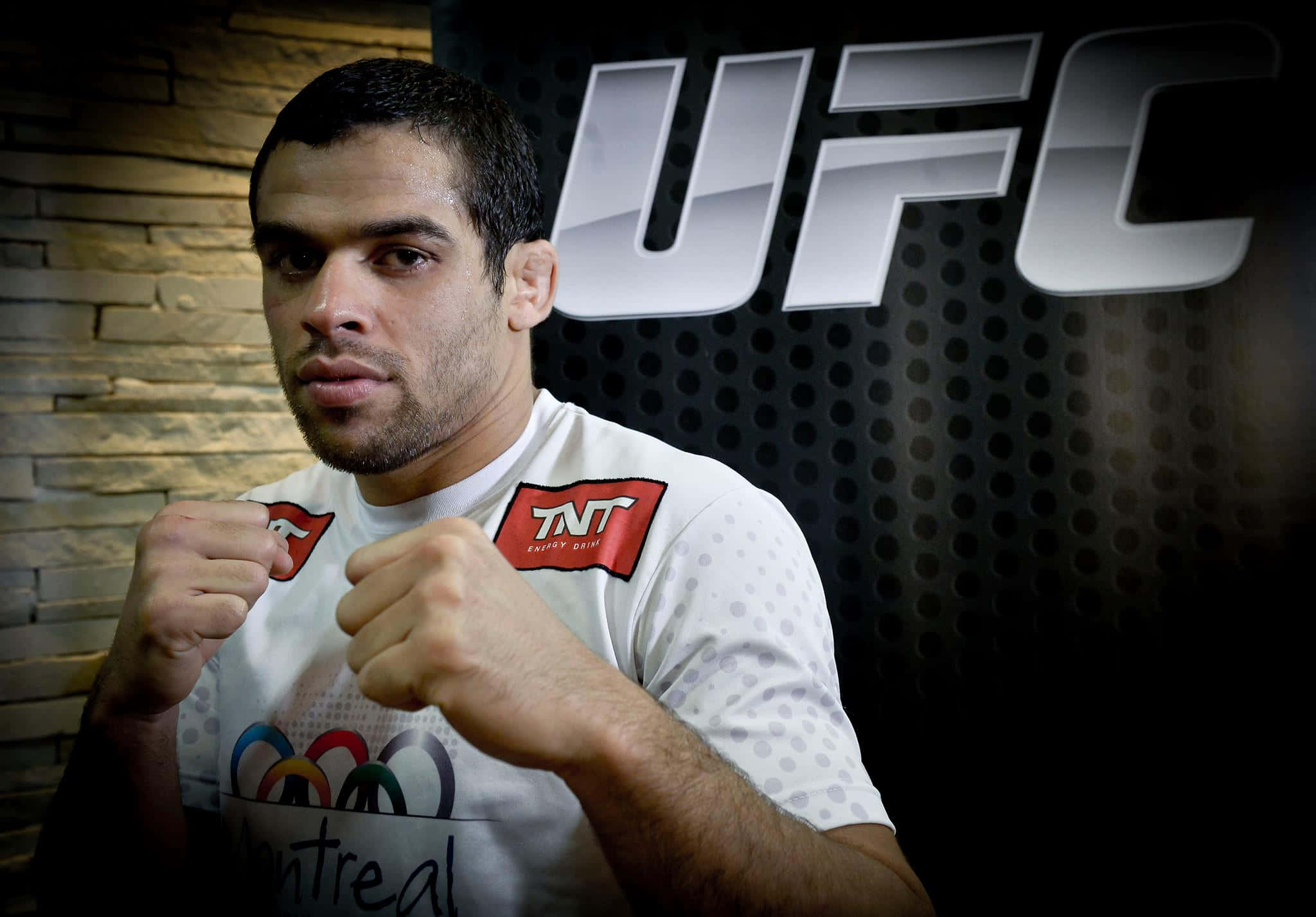 巴西綜合格鬥選手Renan Barão於黑色UFC背景前準備對戰 壁紙 — 一位專注的UFC拳手身穿白色襯衫，在深色穿孔牆前舉起拳頭，身後有UFC標誌。