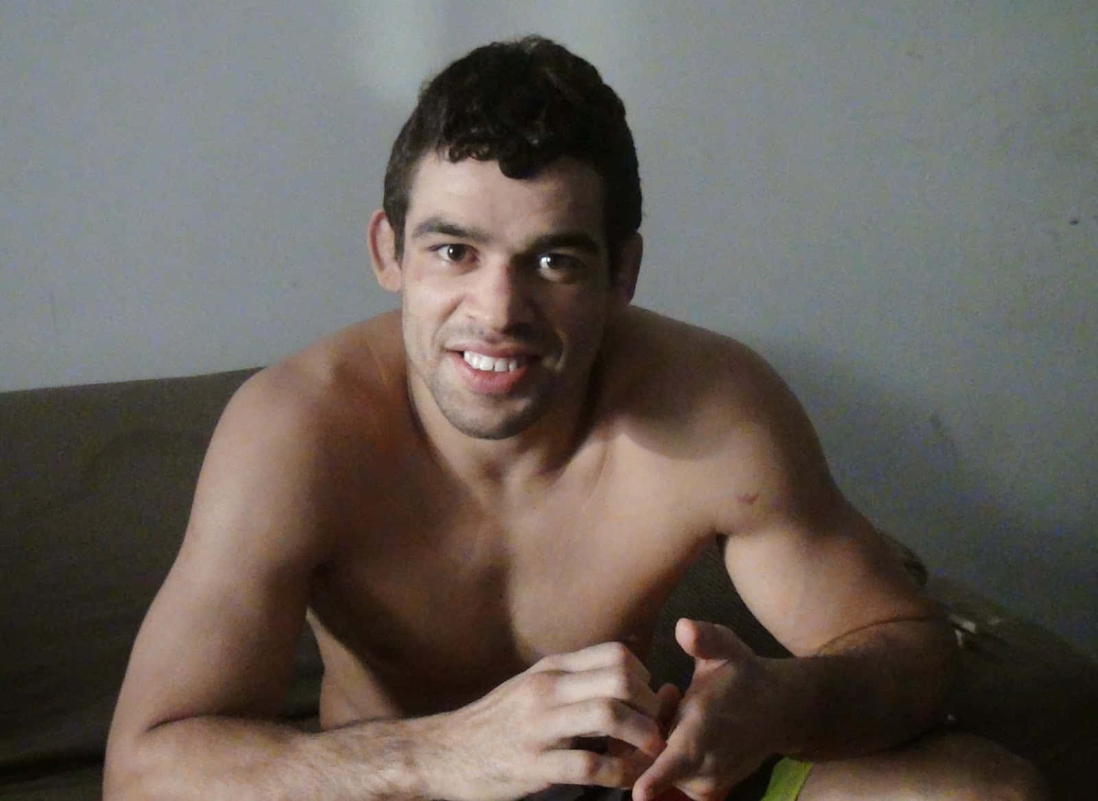 巴西綜合格鬥選手Renan Barão中景微笑坐於棕色沙發 壁紙 — 一位赤膊、肌肉發達、深色頭髮的男子坐在光線昏暗的房間的沙發上，直視鏡頭微笑。