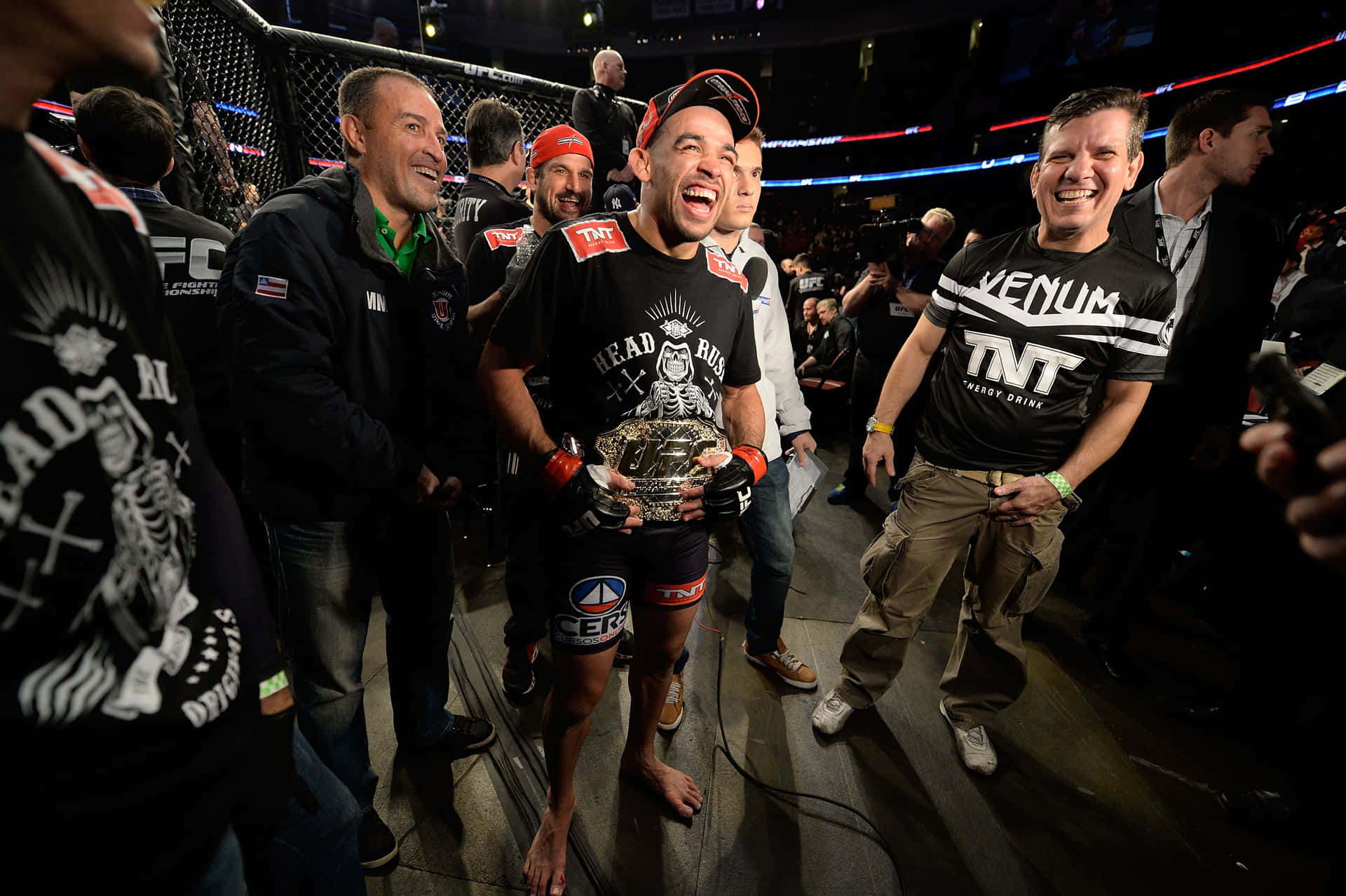 巴西綜合格鬥選手雷南·巴羅手持UFC腰帶 壁紙 — 一位勝利的MMA拳手在燈光明亮的八角籠中，被歡呼的團隊成員包圍，高興地笑著舉起冠軍腰帶。