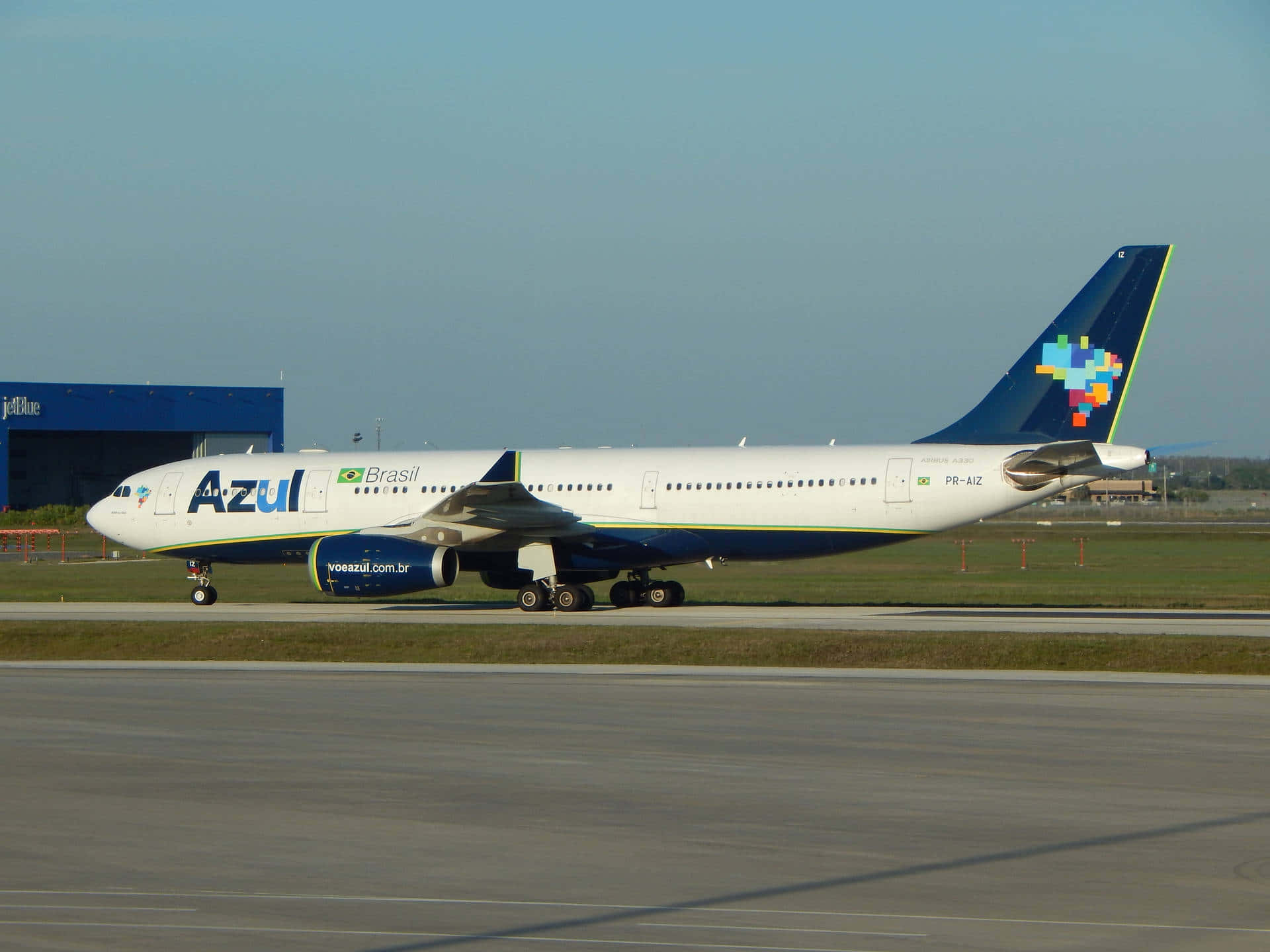 巴西航空A330在滑行道待命起飛 壁紙 — 一架Azul Brasil的空中巴士A330在陽光普照的機場停機坪上滑行，背景是晴朗的藍天和遠處的機庫。