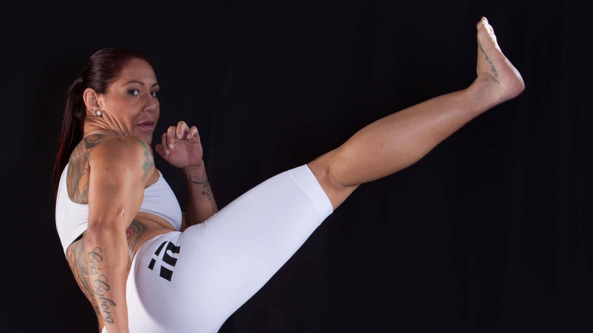 巴西裔美國MMA選手Cris Cyborg寫真照 壁紙 — 一位有紋身的女性穿著白色運動服，在黑色背景下執行高踢腿動作。