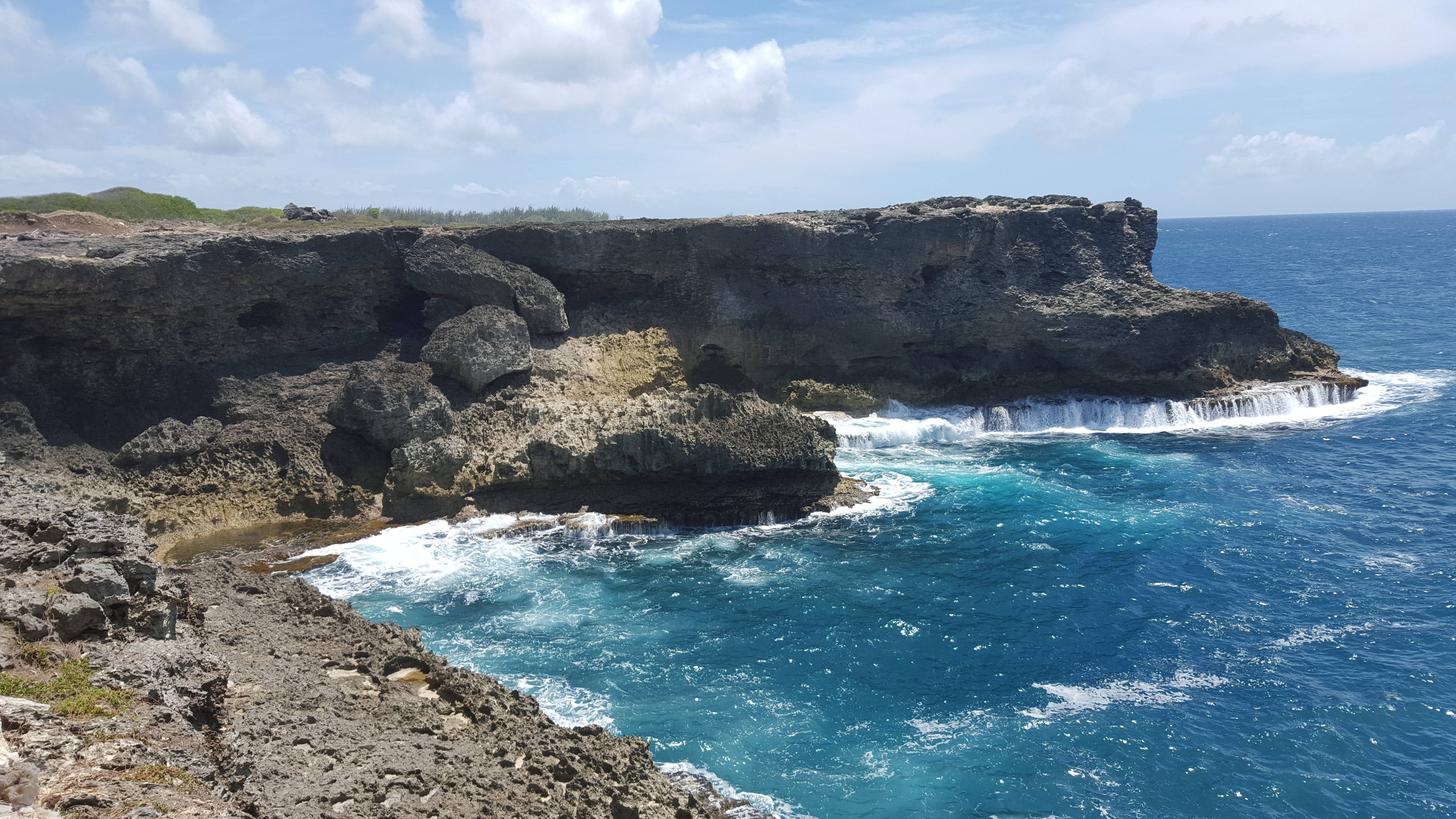 巴貝多島海景 壁紙 — 崎嶇的懸崖陡峭地插入碧綠的海水中，海浪在明亮的藍天下拍打著岩石海岸。
