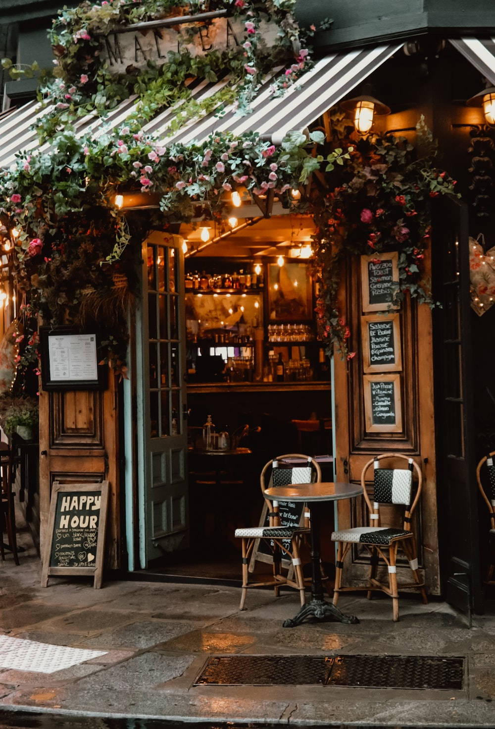 巴黎 Le Malabar 咖啡館酒吧 壁紙 — 迷人的復古咖啡館入口，裝飾著茂盛的花朵和溫暖的燈串，邀請顧客進入其舒適的室內。