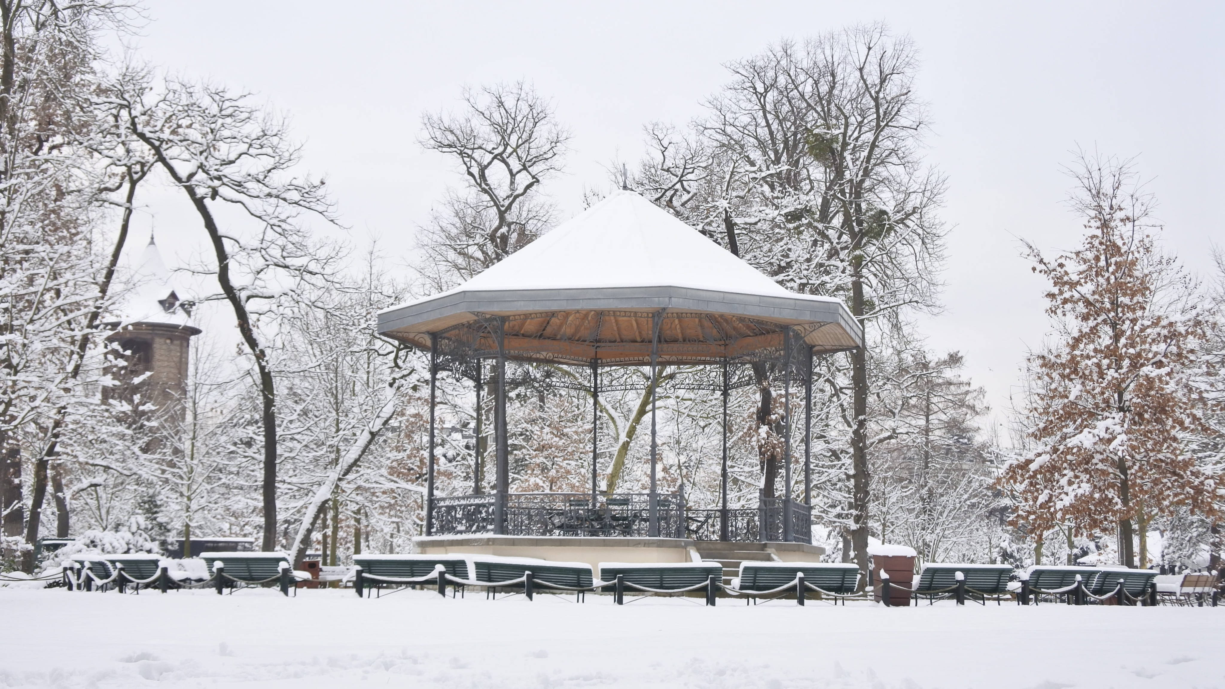 巴黎園林寒意 壁紙 — 一個被雪覆蓋的音樂亭，位於冬季公園中，周圍是光禿禿的樹木和長椅，天空多雲。