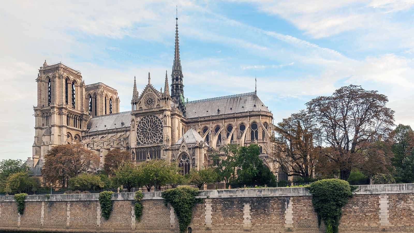 Gothic Architecture 桌布 壁紙 — 巴黎聖母院在部分多雲的天空下巍然聳立，秋天的樹木襯托出其標誌性的哥德式建築。