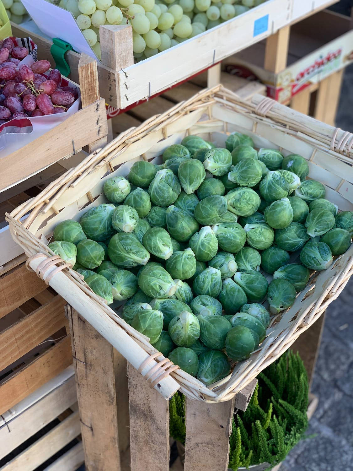 市場新鮮捲心菜展示 壁紙 — 新鮮的綠色抱子甘藍堆放在一個編織籃子裡，在陽光明媚的戶外市場攤位上，附近有木箱和葡萄。