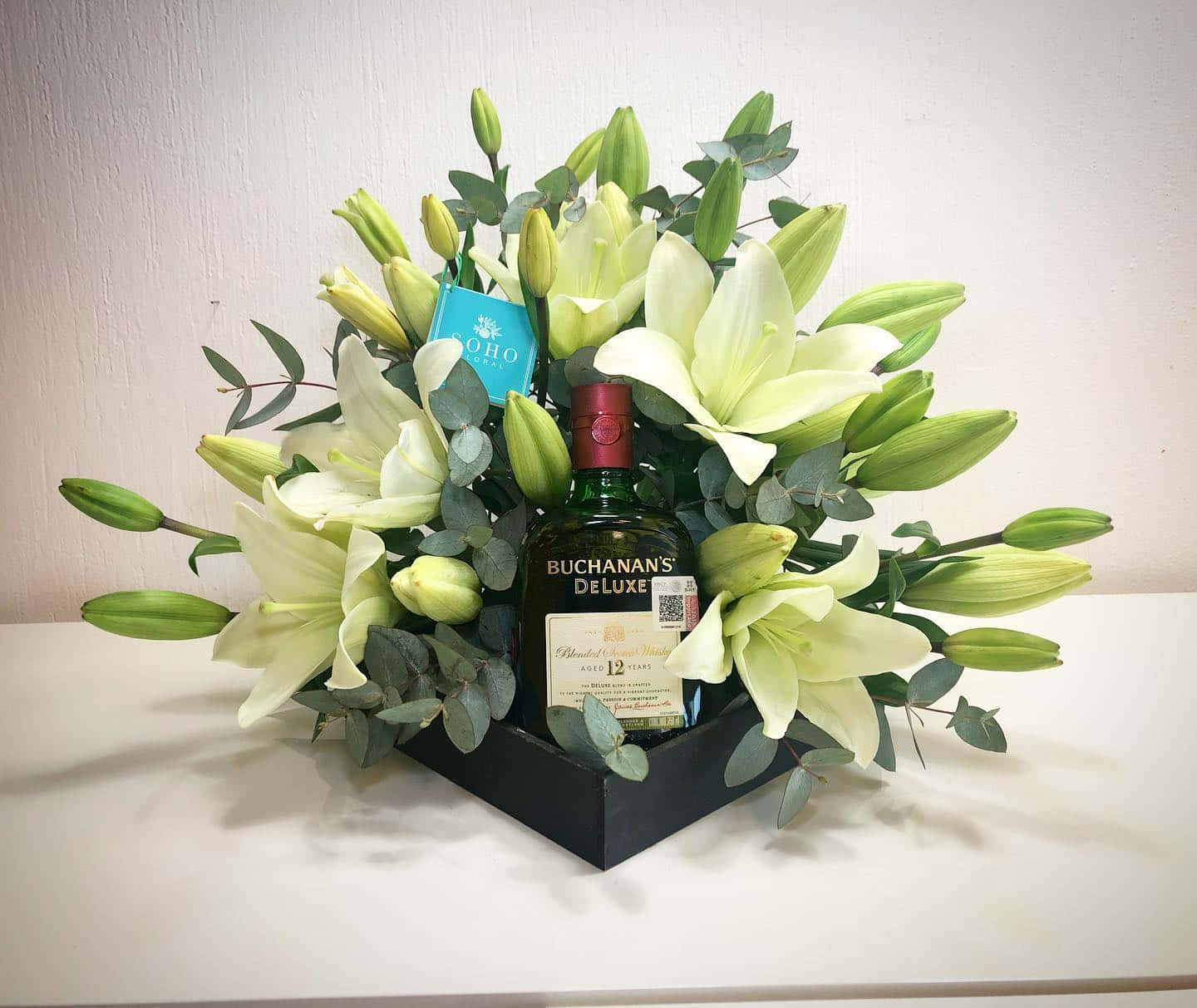 威士忌瓶被白百合和綠色植物環繞，放在黑色禮盒中。