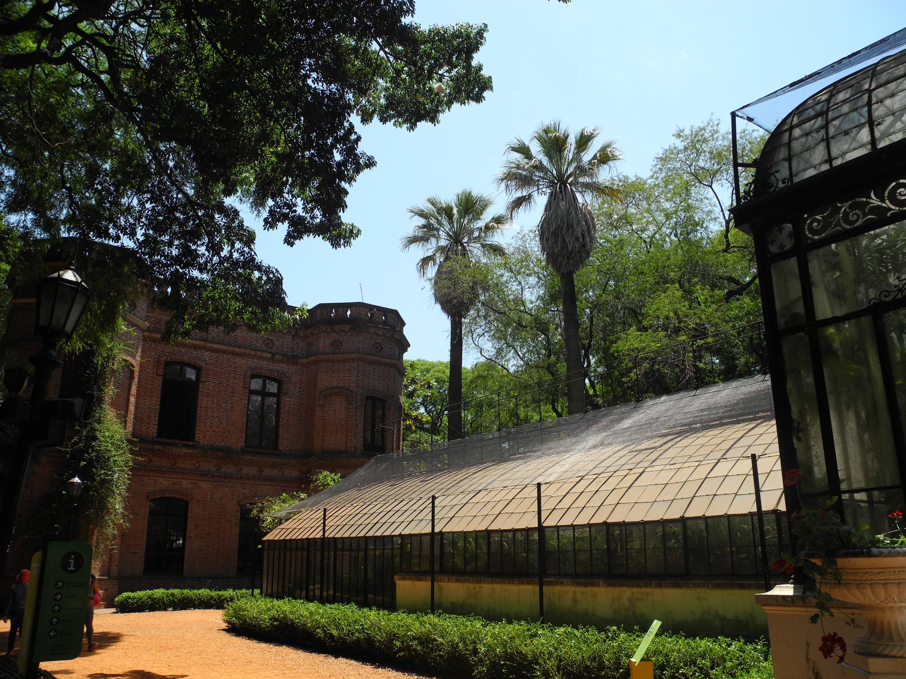 布宜諾斯艾利斯卡洛斯·薩斯植物園 壁紙 — 陽光普照的花園場景，設有紅磚建築、高大的棕櫚樹和玻璃溫室，在晴朗的藍天下。