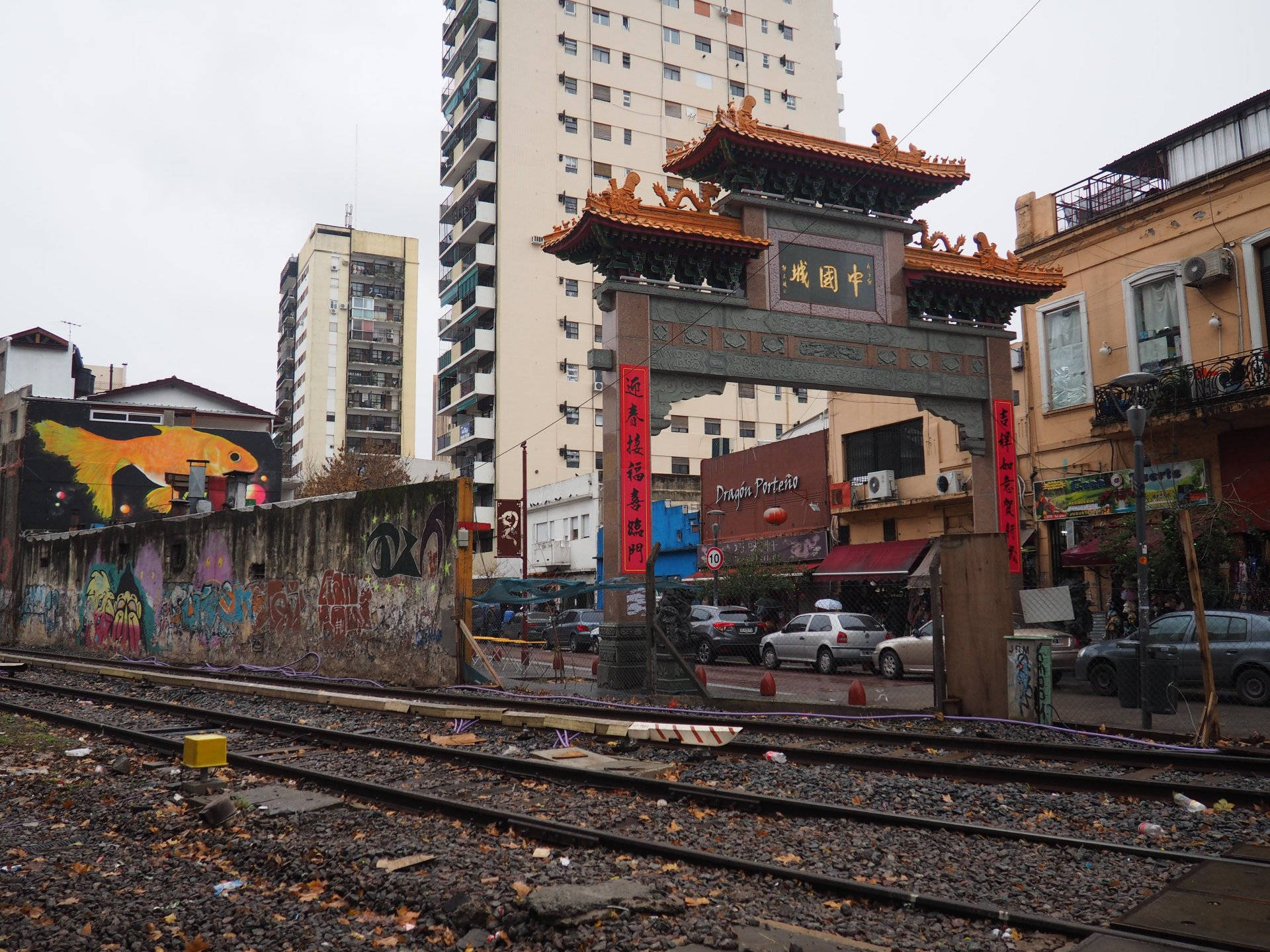 布宜諾斯艾利斯唐人街旁鐵路 壁紙 — 一座傳統中式牌坊矗立於城市衰敗之中，前景是塗鴉牆壁和火車軌道，天空陰沉。