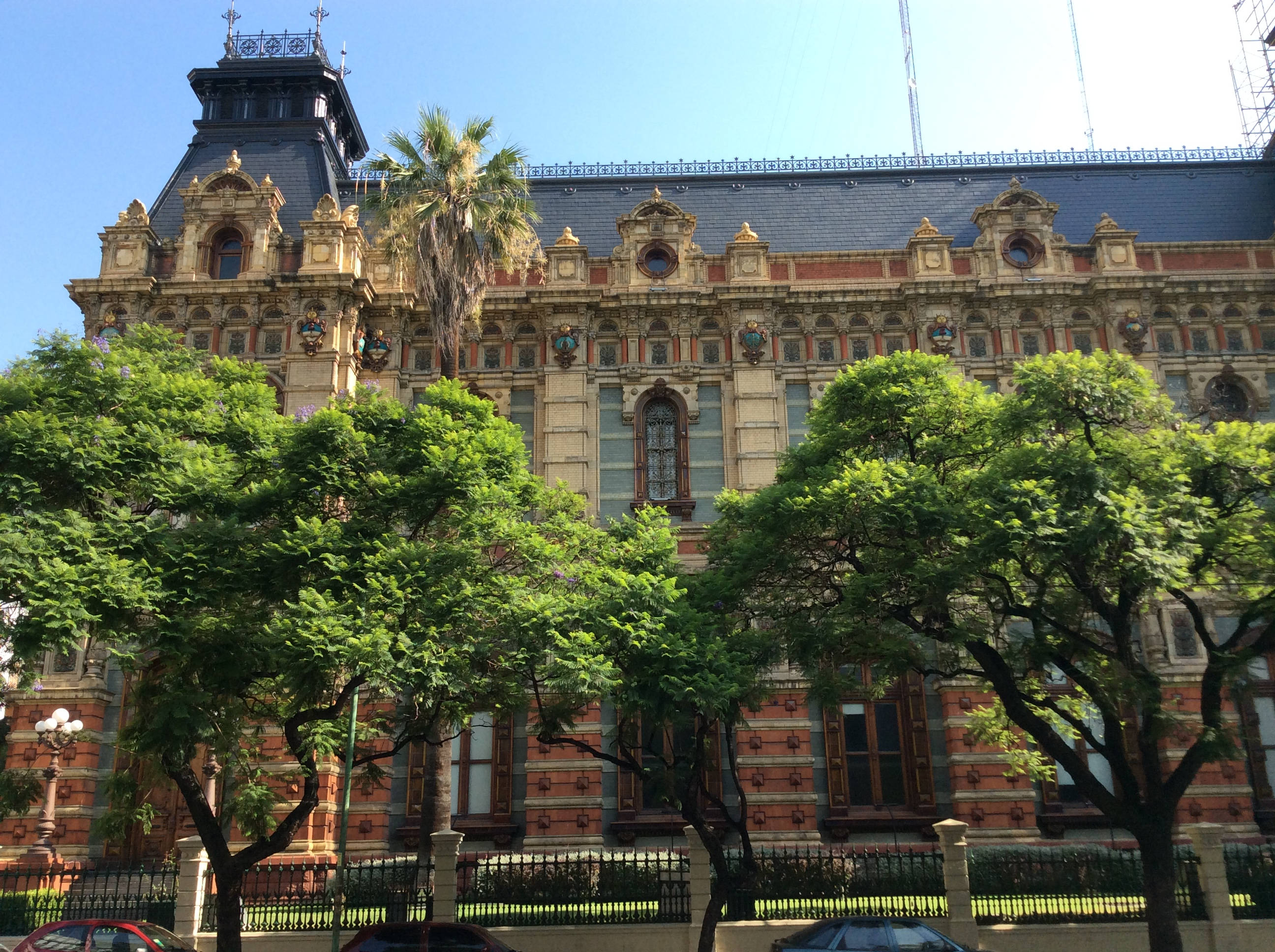 布宜諾斯艾利斯水公司宮殿 壁紙 — 一座宏偉華麗的歷史建築，以紅磚和金色點綴，矗立在茂密的綠樹之後，背景是清澈的藍天。
