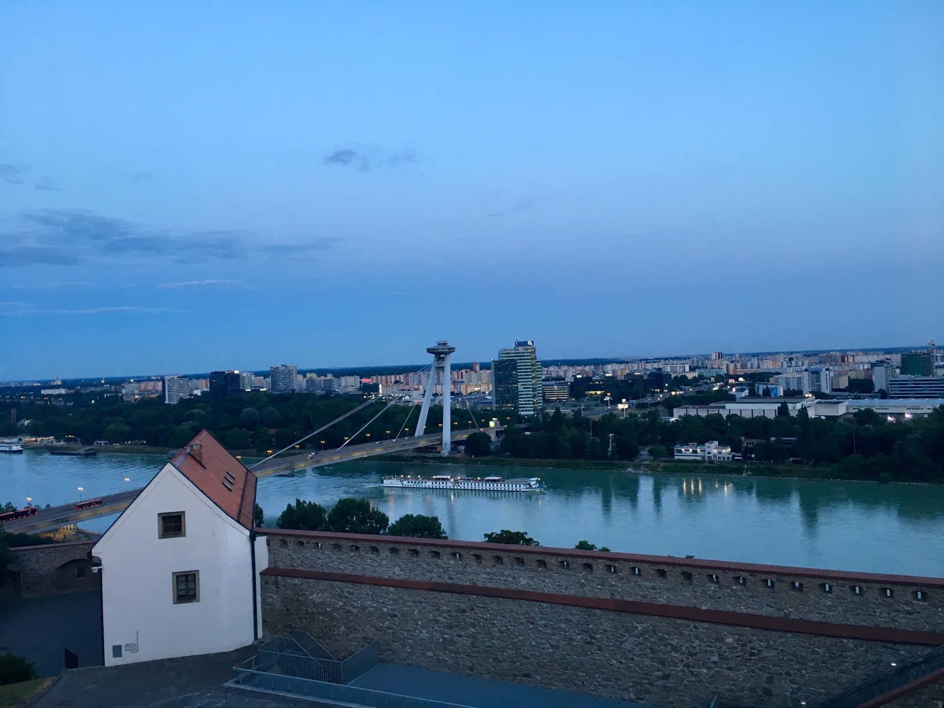 布拉提斯拉瓦城堡俯瞰城市 壁紙 — 黃昏城市景觀，一艘河上遊船在現代懸索橋下滑行，暮色籠罩城市天際線。