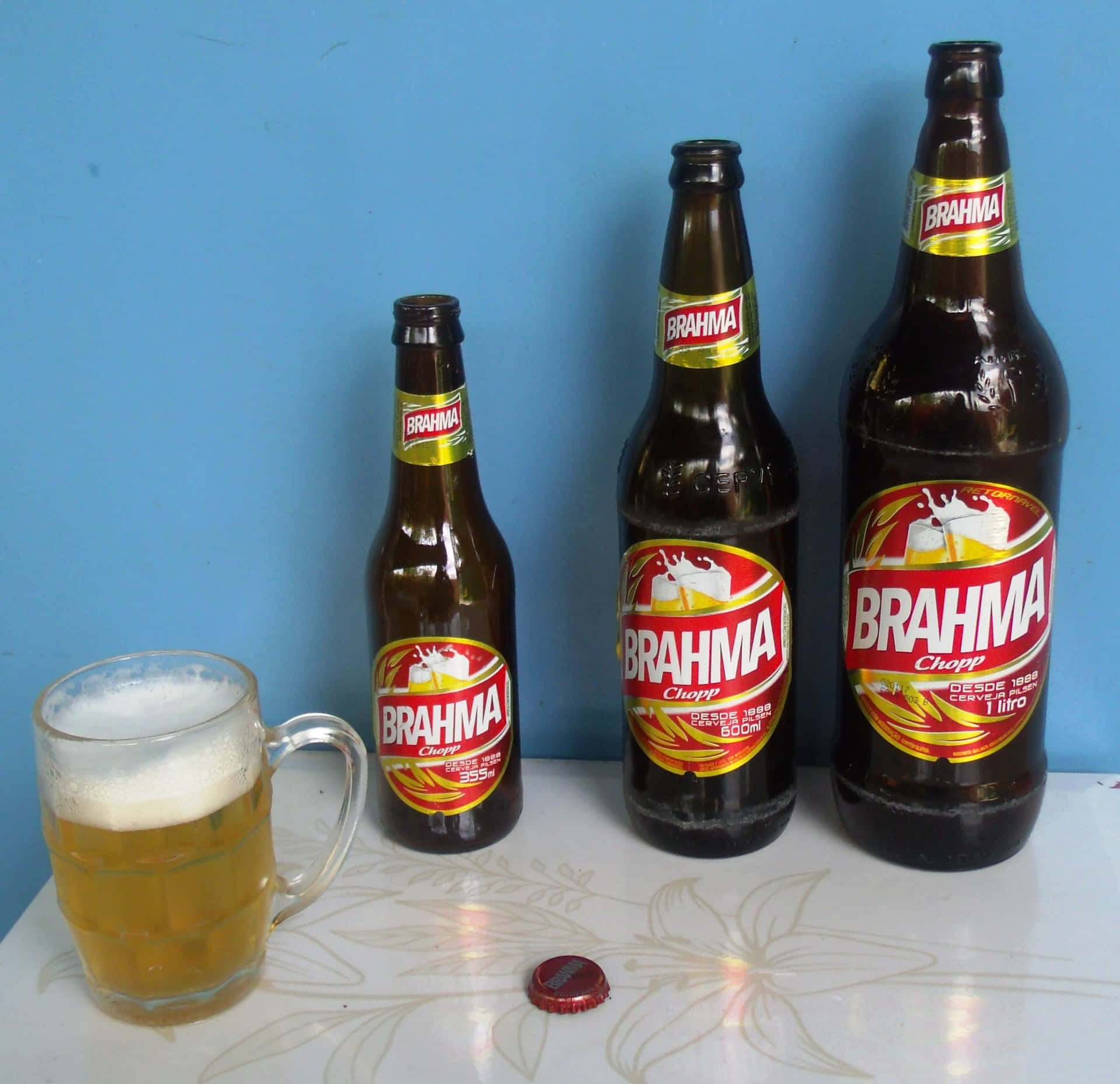 三瓶 Brahma 啤酒和一個裝滿啤酒的馬克杯放在有圖案的桌子上。