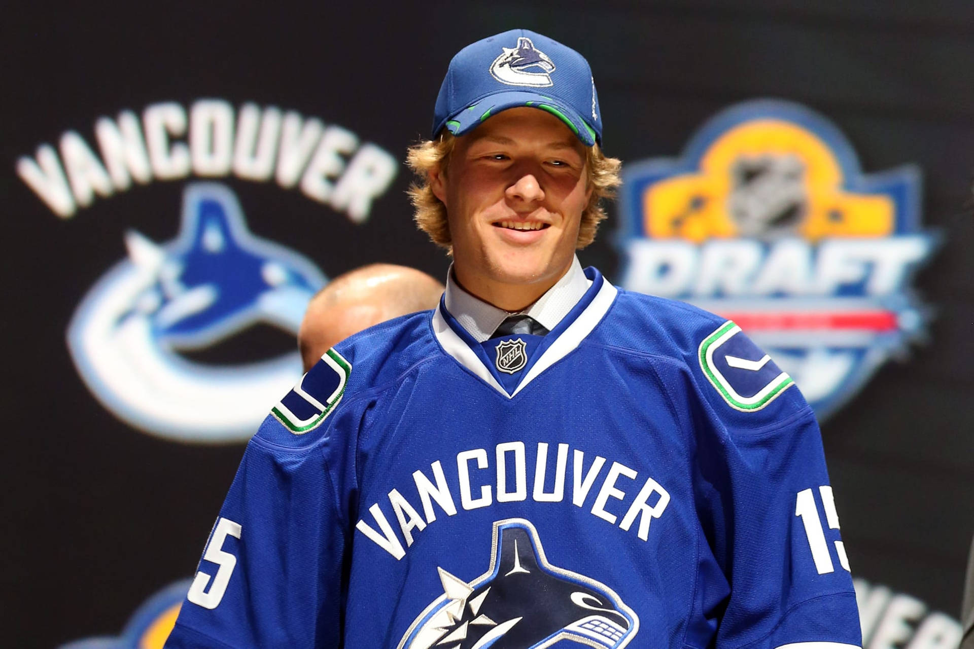 布羅克·博瑟2015年NHL選秀第23順位 壁紙 — 一名身穿溫哥華加人隊藍色球衣的年輕冰球運動員在選秀活動中對著鏡頭微笑。
