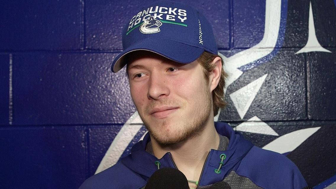 布羅克·博瑟國家冰球聯盟訪問 壁紙 — 一名戴著 Canucks 帽子和藍色連帽衫的曲棍球運動員，在帶有球隊標誌的藍色磚牆前對著麥克風說話，微微一笑。