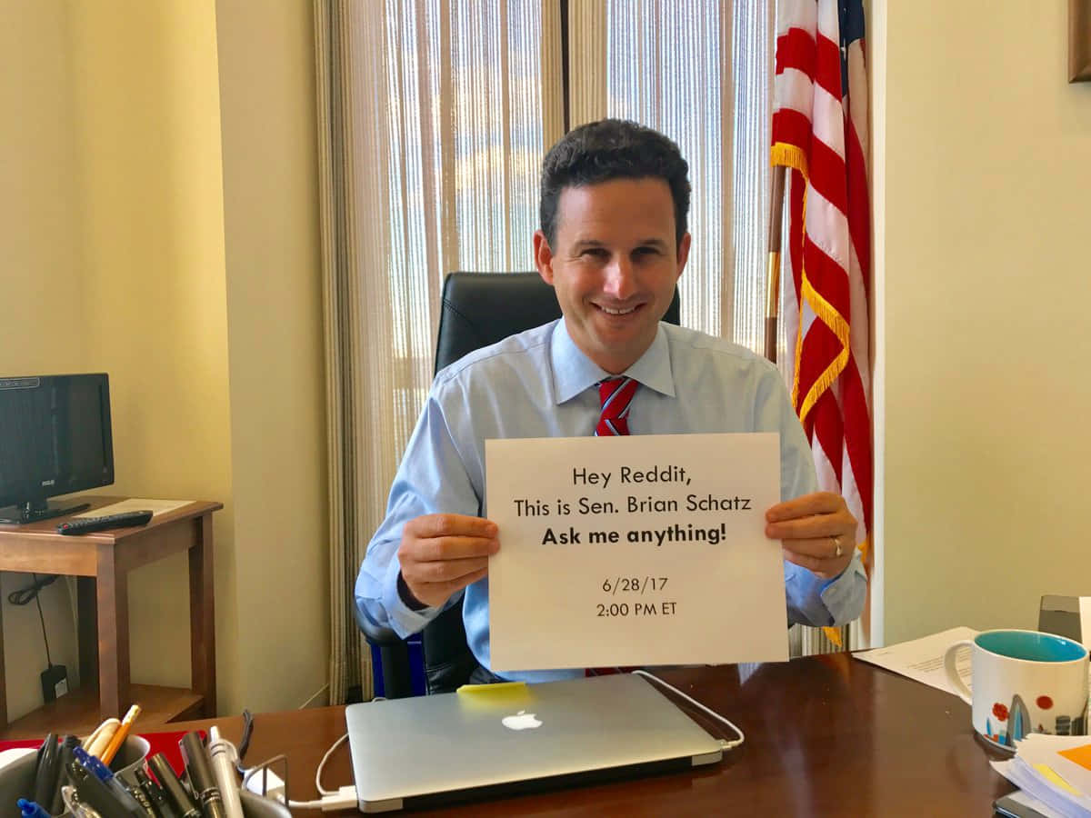 布萊恩·沙茲參加 Reddit AMA 壁紙 — 參議員Brian Schatz微笑著，在他的辦公室裡拿著一個邀請Reddit問題的牌子，身後有美國國旗。