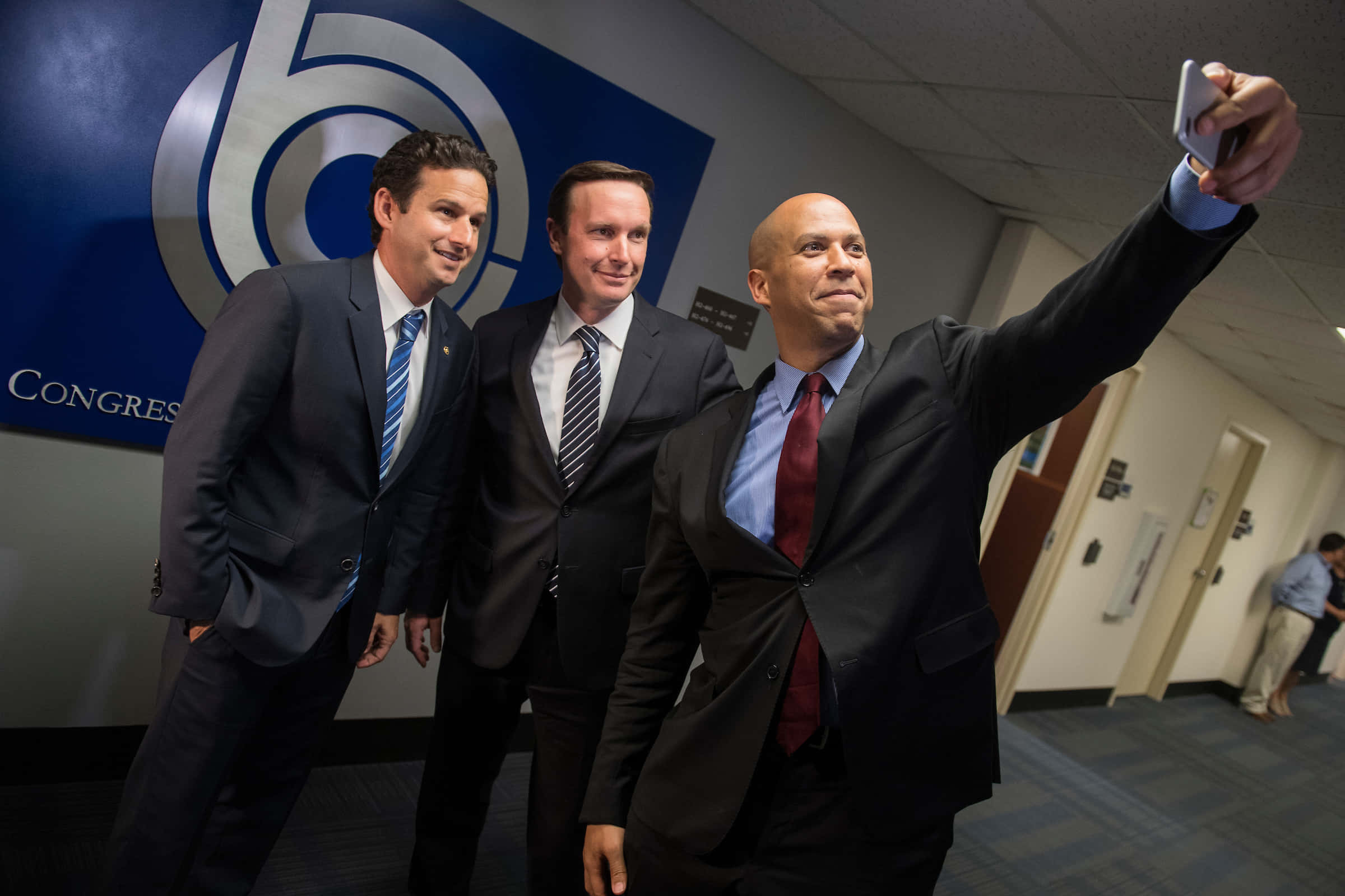 布萊恩·沙茲與科里·布克自拍合影 壁紙 — 三名身穿西裝的男子在國會走廊中以藍色標誌牆為背景自拍。