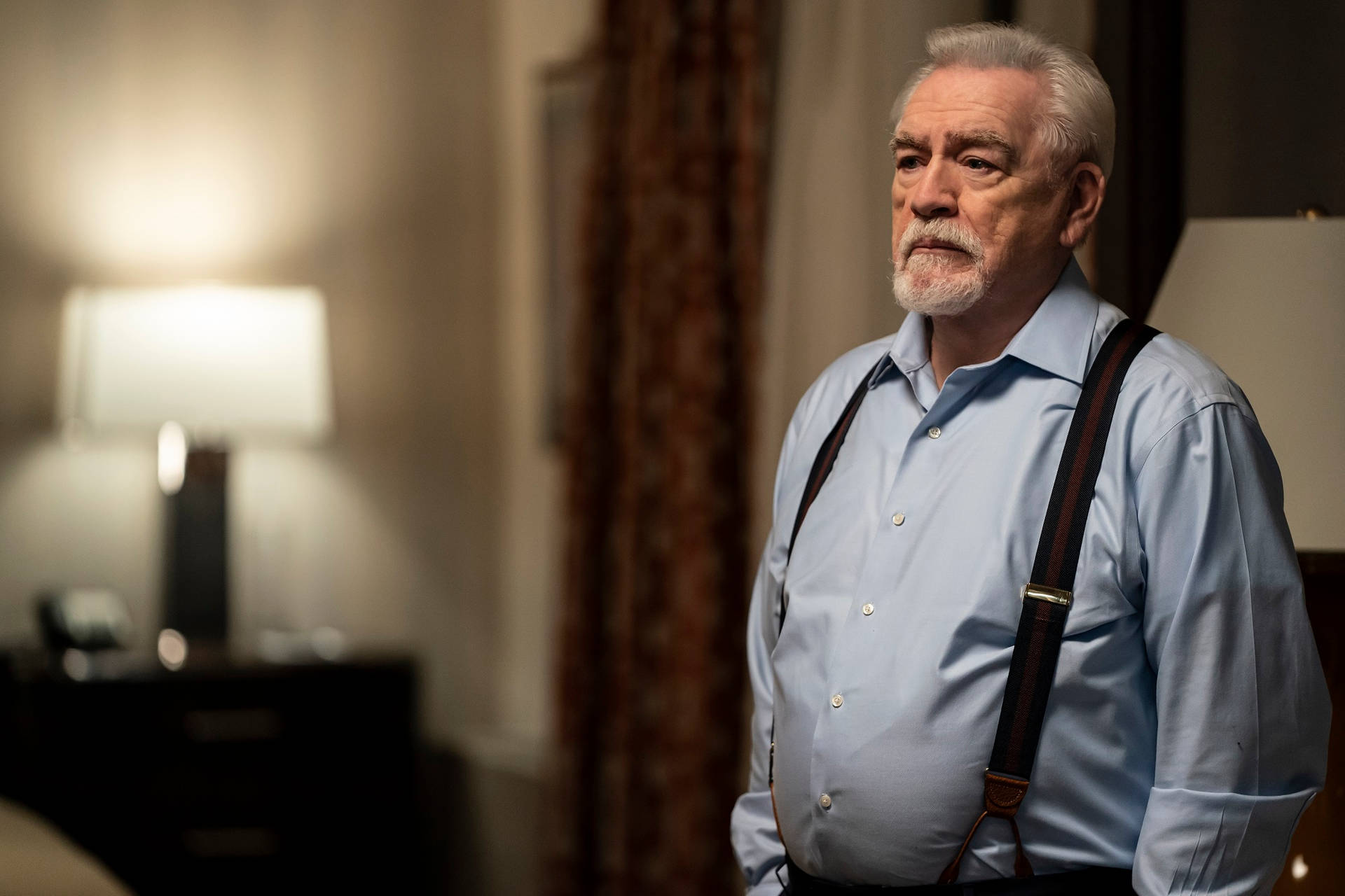一位留著白鬍子的成熟男士在光線溫暖的房間裡穿著吊帶褲和藍色襯衫。
