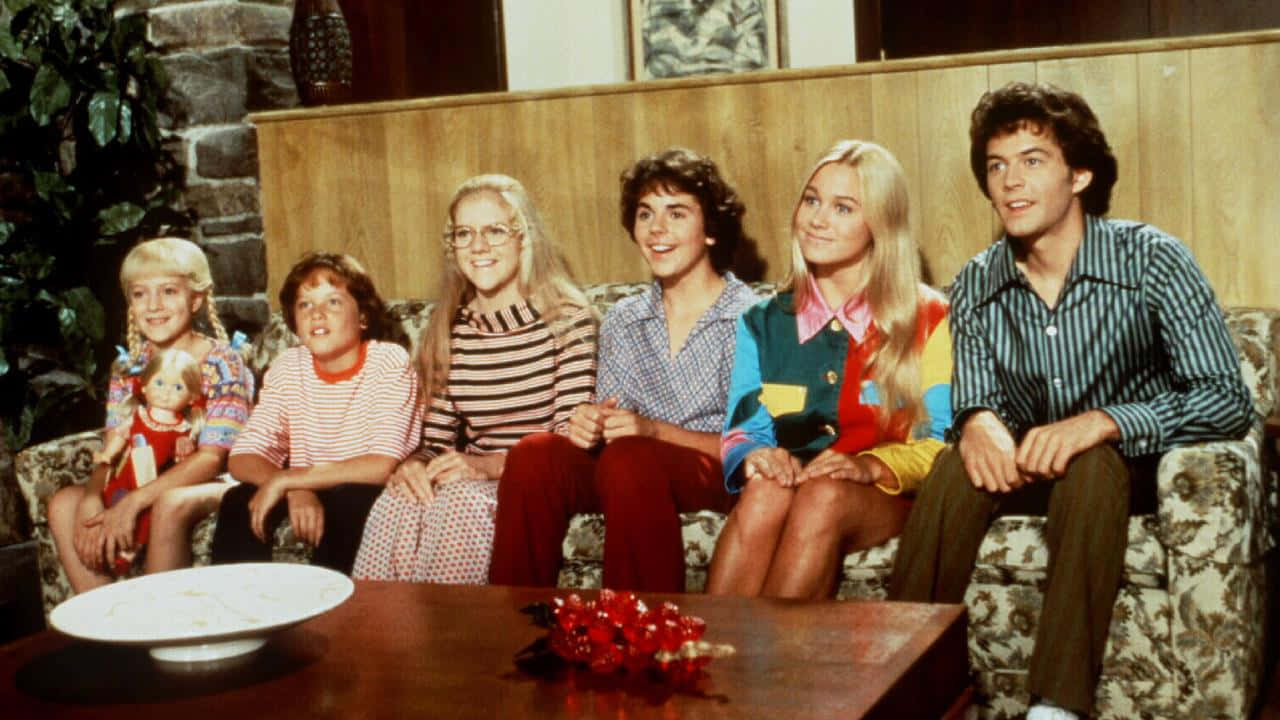 六個穿著1970年代服裝的人坐在客廳的花卉沙發上。