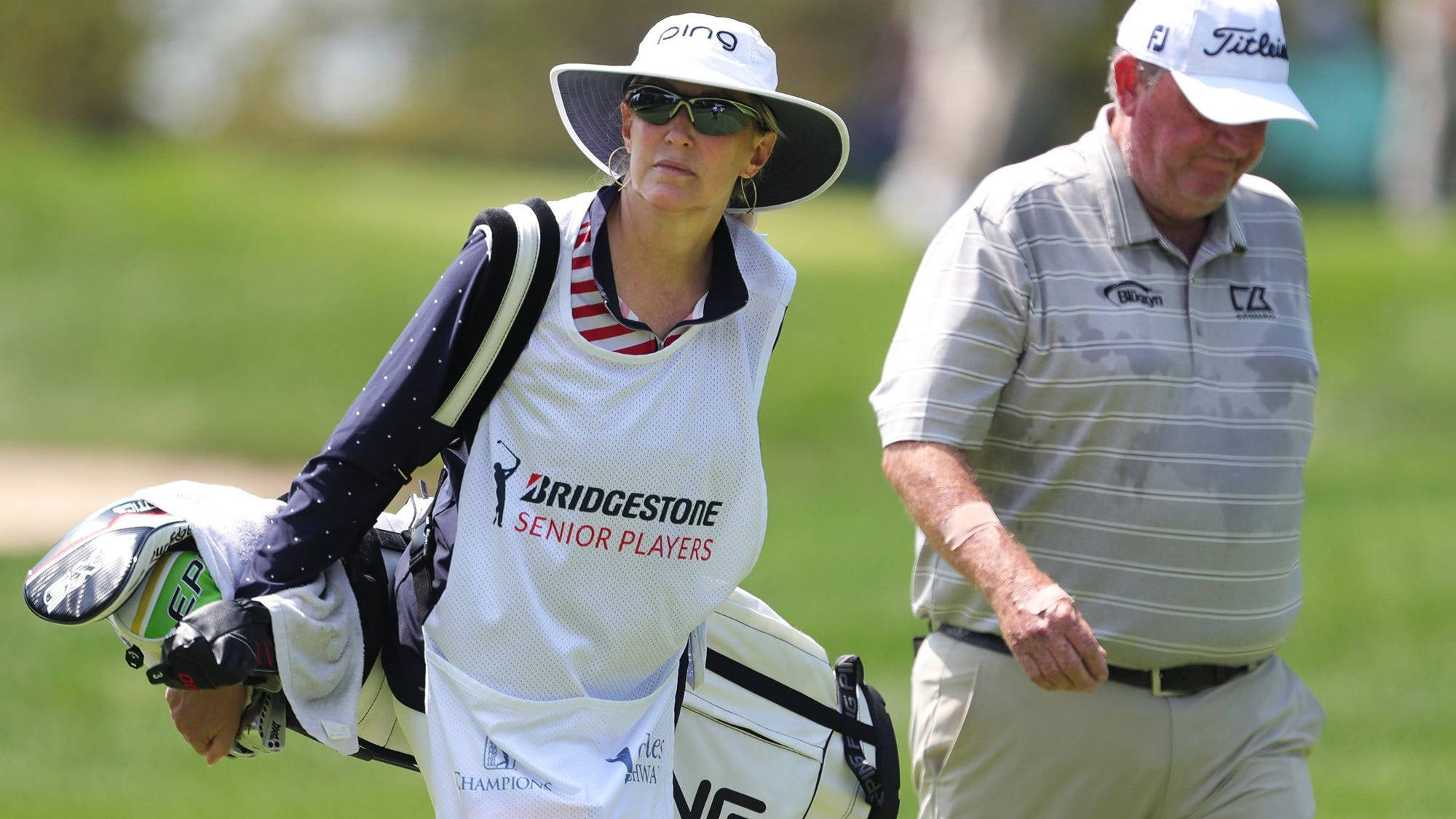 布蘭達與馬克·卡爾卡維奇 壁紙 — 一位穿著Bridgestone Senior Players背心的女高爾夫球手，在陽光明媚的高爾夫球場上，背著球袋走在一位年長男高爾夫球手旁邊。