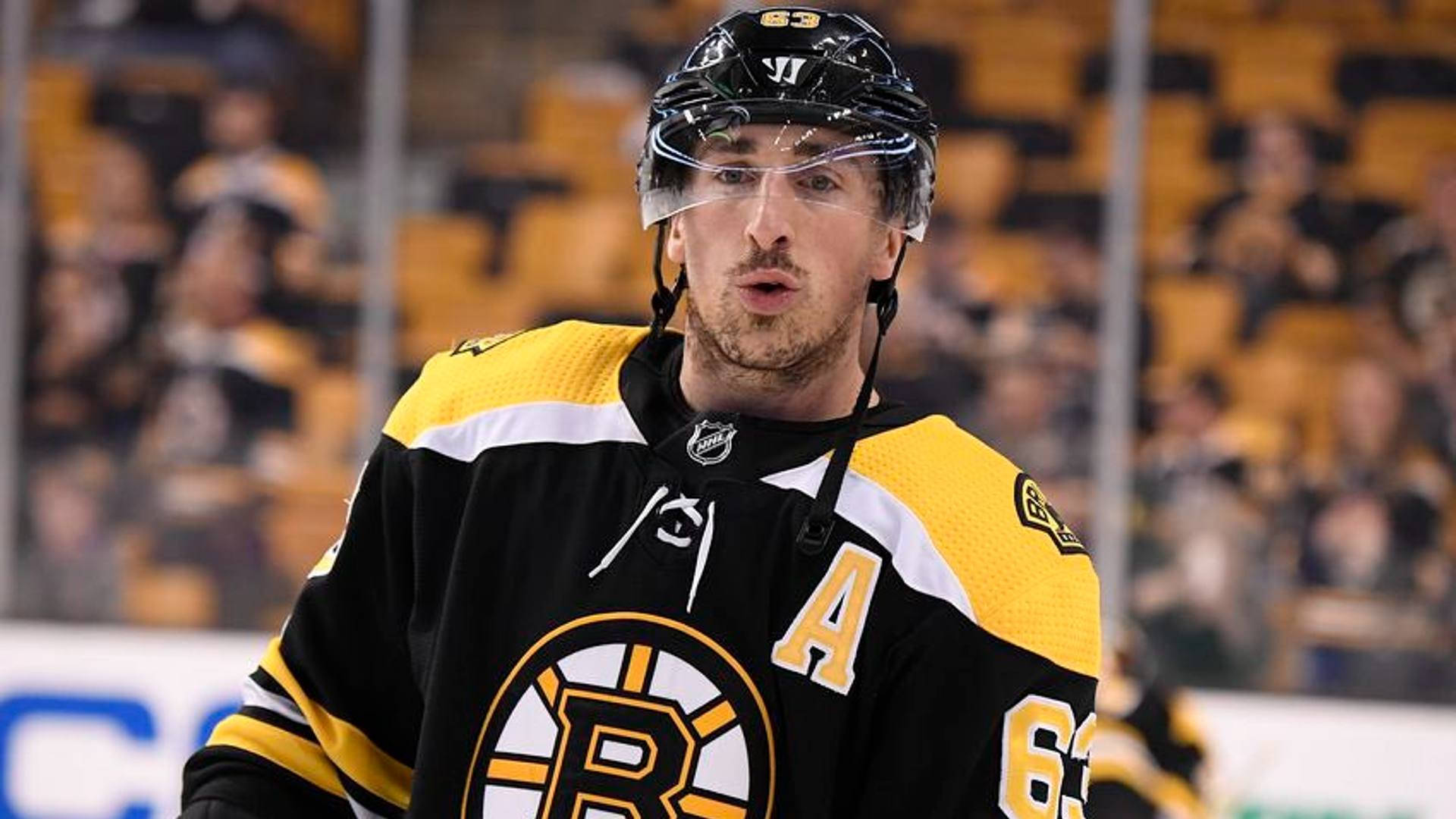 布雷德·馬奇安德NHL 壁紙 — 一名身穿黑色和金色波士頓棕熊隊球衣（號碼63，字母A）的冰球運動員在比賽中站在冰上，身後黃色座位上的球迷模糊不清。