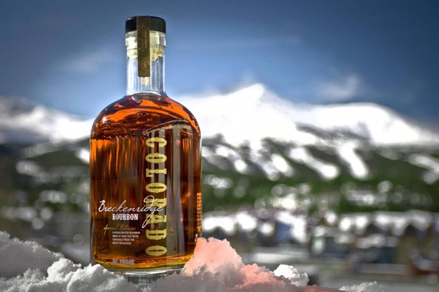 布雷肯里奇蒸餾廠波本威士忌山景 壁紙 — 一瓶 Breckenridge Colorado 波本威士忌放在新雪中，背景是白雪覆蓋的山脈和清澈的藍天。