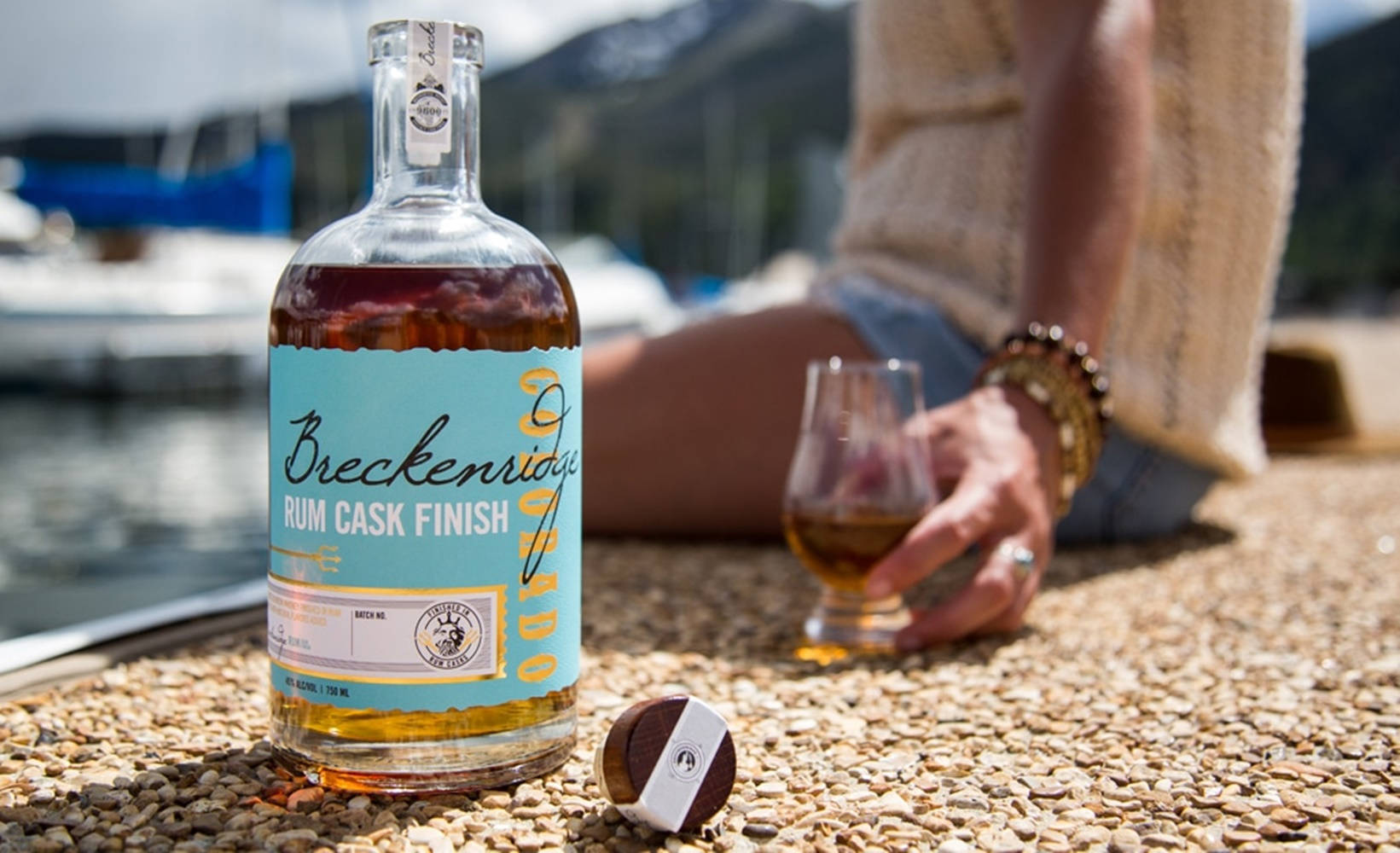 布雷肯里奇蒸餾廠波本朗姆桶釀威士忌 壁紙 — 一瓶 Breckenridge Rum Cask Finish 威士忌放在鵝卵石上，旁邊有一個人拿著玻璃杯，背景是水、船隻和山脈。