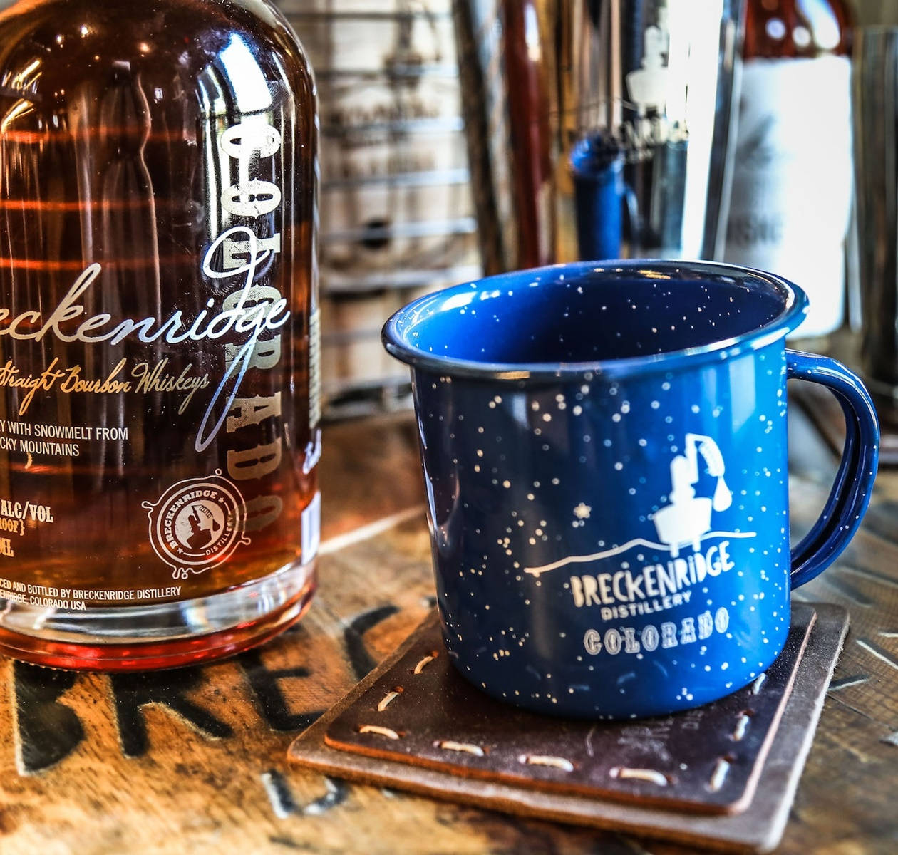 布雷肯里奇蒸餾廠烈酒與藍色商品杯 壁紙 — 一個帶有 Breckenridge Distillery 標誌的藍色斑點馬克杯放在一瓶波本威士忌旁邊，置於鄉村風格的木製吧檯上。