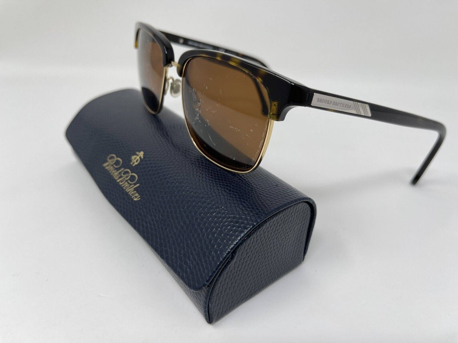 布魯克斯兄弟男士眼鏡 壁紙 — 經典玳瑁太陽眼鏡搭配金色點綴，優雅地放置在帶有紋理的深藍色Brooks Brothers眼鏡盒上，背景為純白色。