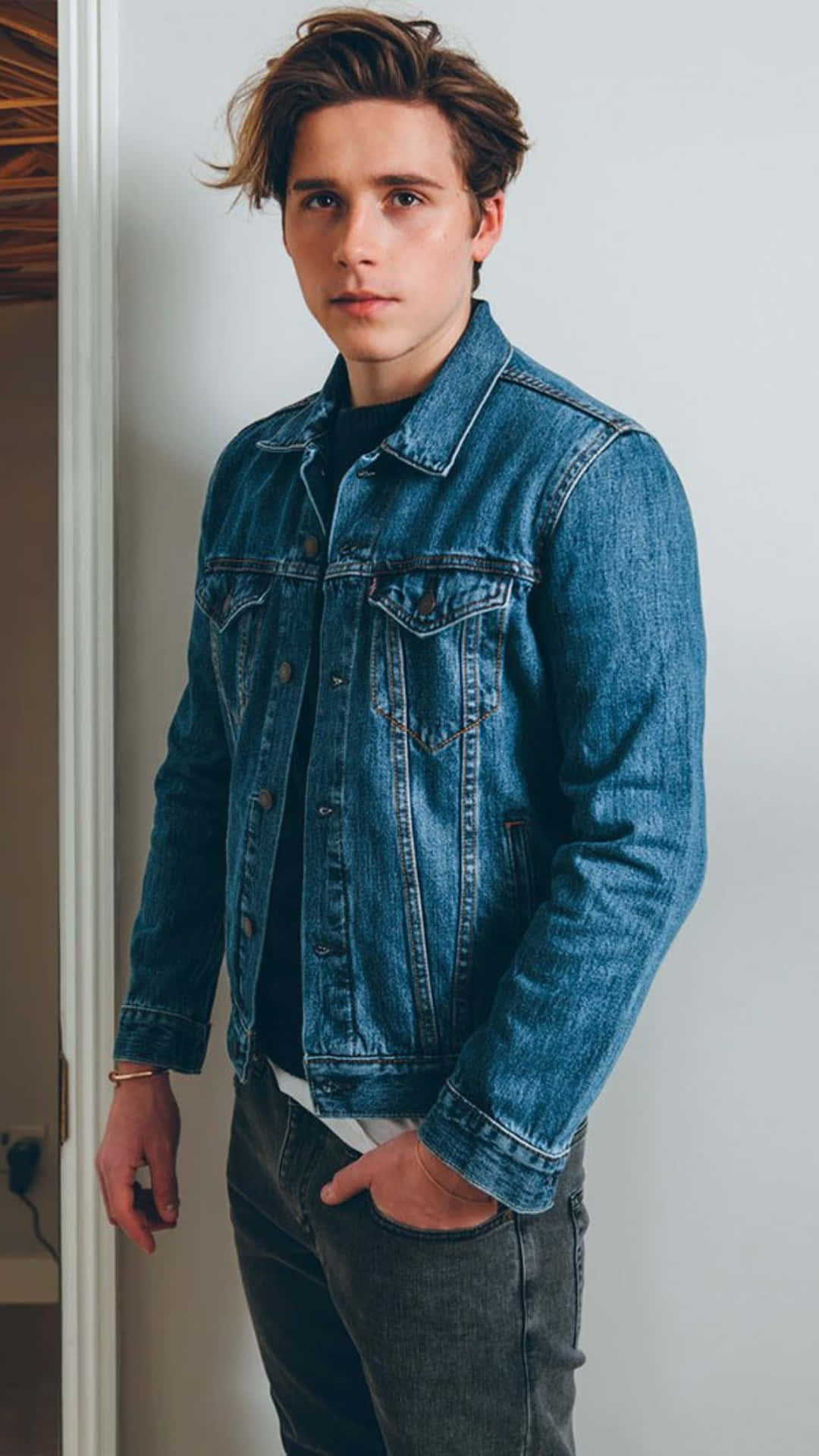 布魯克林·貝克漢姆休閒牛仔風格 壁紙 — 一位年輕男子，棕色亂髮，身穿經典藍色牛仔外套，內搭黑色襯衫和深色牛仔褲，在室內靠著白色牆壁。