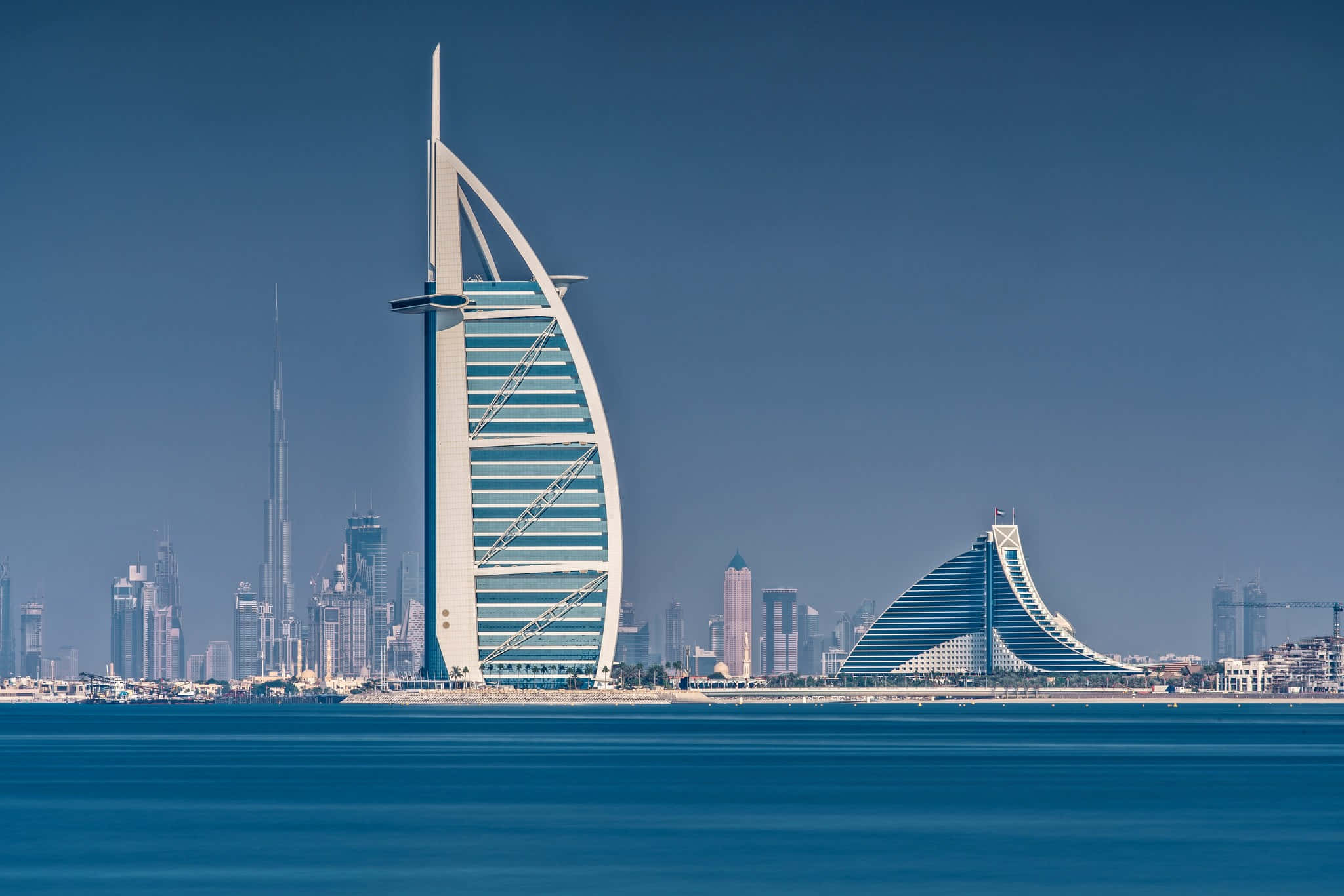 Burj Khalifa 桌布 壁紙 — 帆船酒店（Burj Al Arab）矗立在清澈的藍天下，平靜的水面映照出其標誌性的帆船形狀。