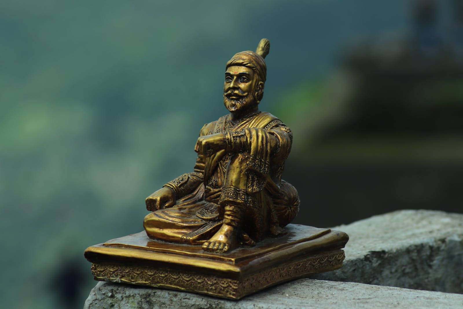 金色雕像，一個坐著嘅鬍鬚男，穿著傳統衫，喺石底座上。