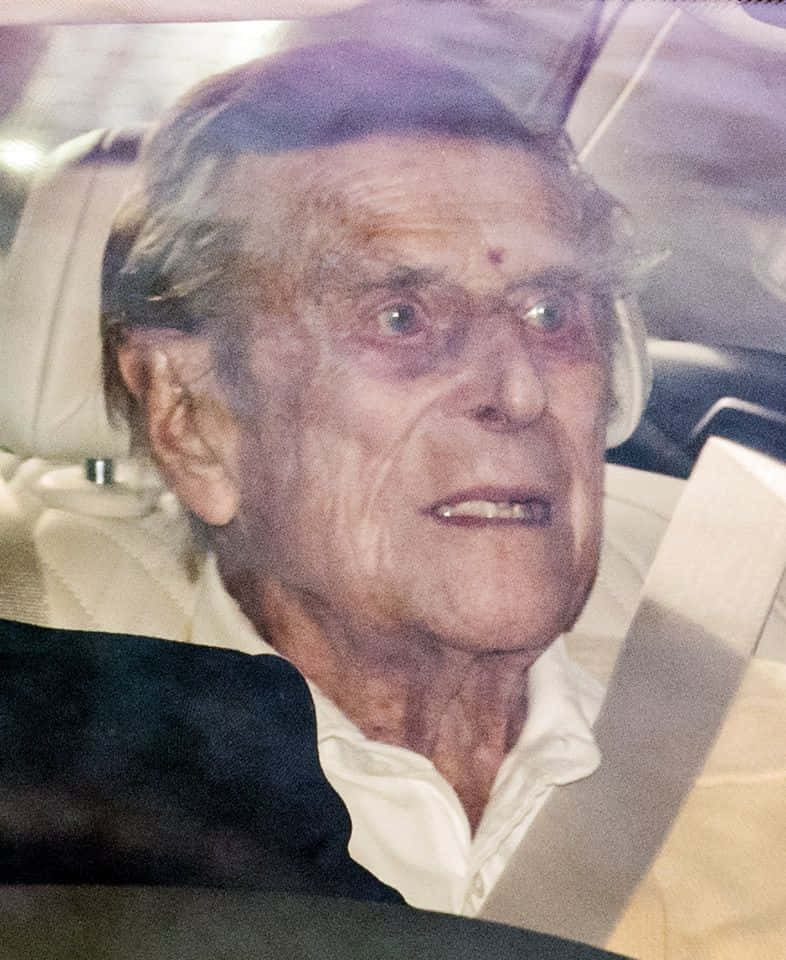 一位灰髮、額頭有紅點的長者，穿著白恤衫坐在汽車裡。