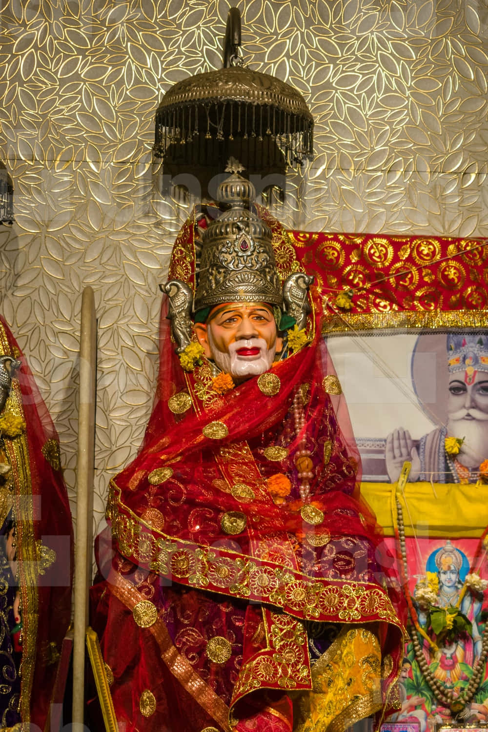 希迪 Sai Baba 的神聖肖像 壁紙 — 一尊裝飾華麗的宗教雕像，披著紅金布料，繡工精緻，頭戴鑲有寶石的皇冠，立於質感豐富的金箔牆前。