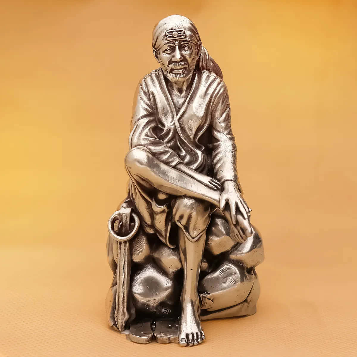 希迪 Sai Baba 銀色雕像肖像 壁紙 — 一個詳細的銀色雕像，描繪一位戴頭巾、持杖的坐姿精神導師，在溫暖漸變背景前擺出冥想姿勢。