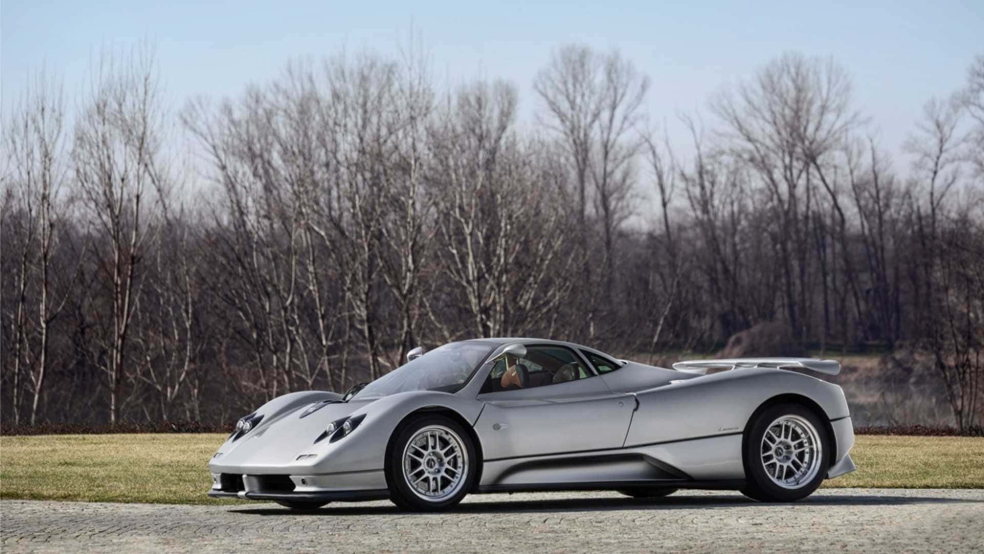 帕加尼Zonda C12 Roadster奢華超跑行駛 壁紙 — 一輛時尚的銀色Pagani超級跑車停在鵝卵石路上，背景是光禿禿的樹木，天空晴朗。
