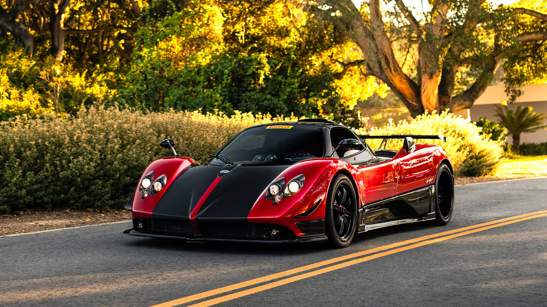 日落時分，一輛紅色和黑色的Pagani Zonda跑車行駛在綠樹成蔭的道路上。