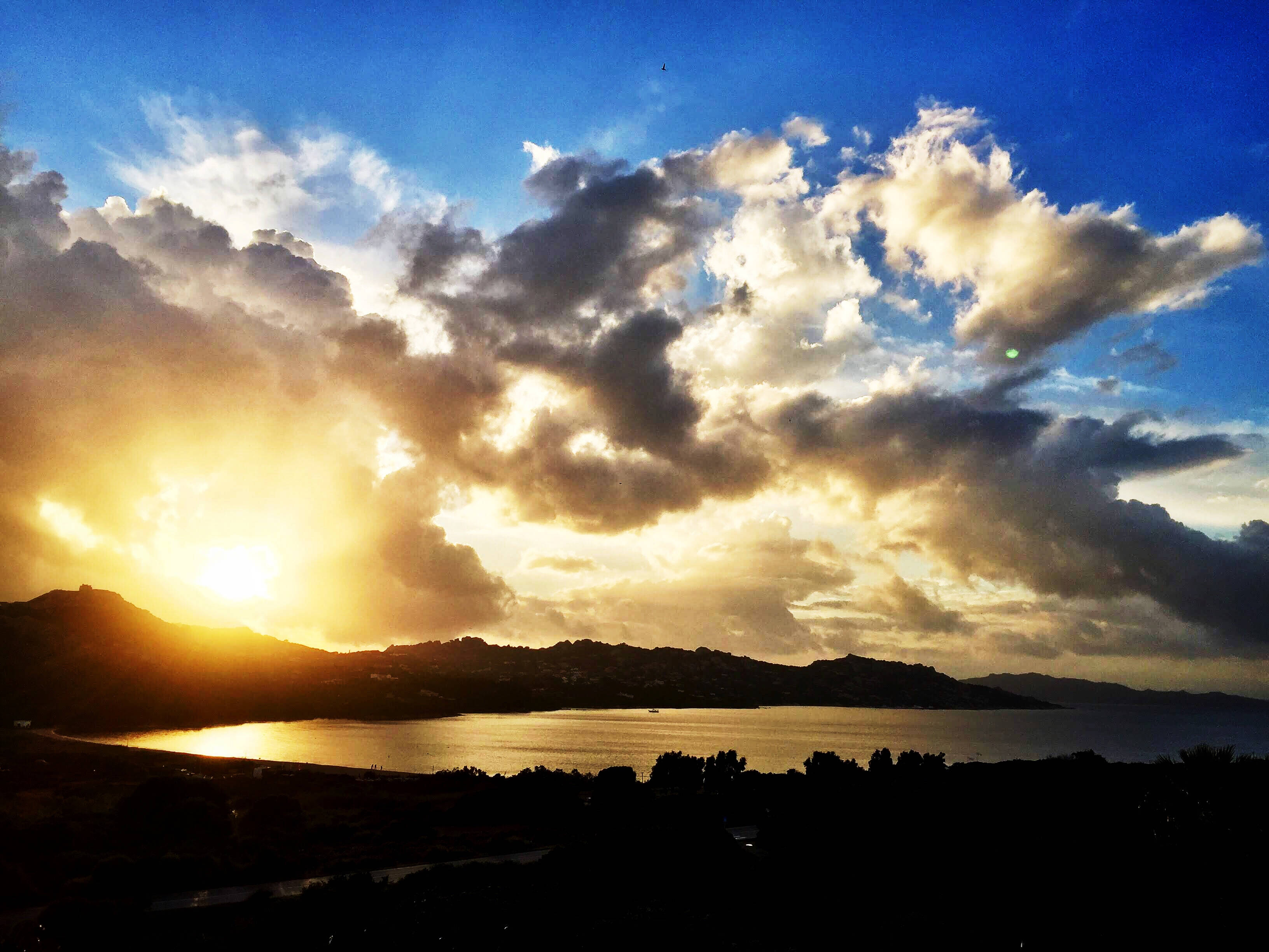 帕勞山巒日落 壁紙 — 夕陽將戲劇性的雲朵沐浴在金色光芒中，映照著輪廓分明的海岸灣和山丘。