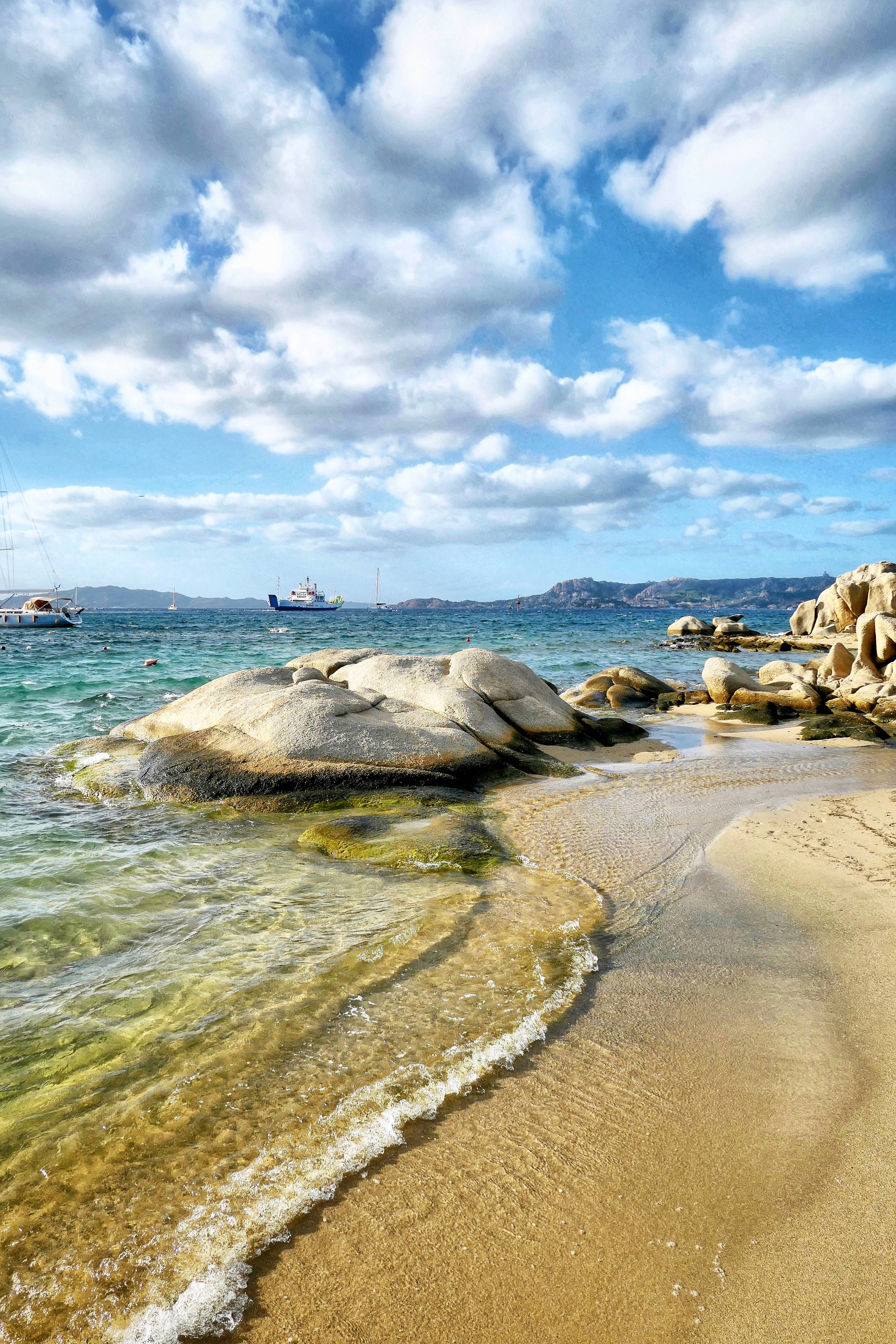 帕勞海浪拍打岩石 壁紙 — 陽光普照的碧綠海浪拍打著金色沙灘，旁邊是風化的岩石，上方是點綴著蓬鬆白雲的明亮藍天。