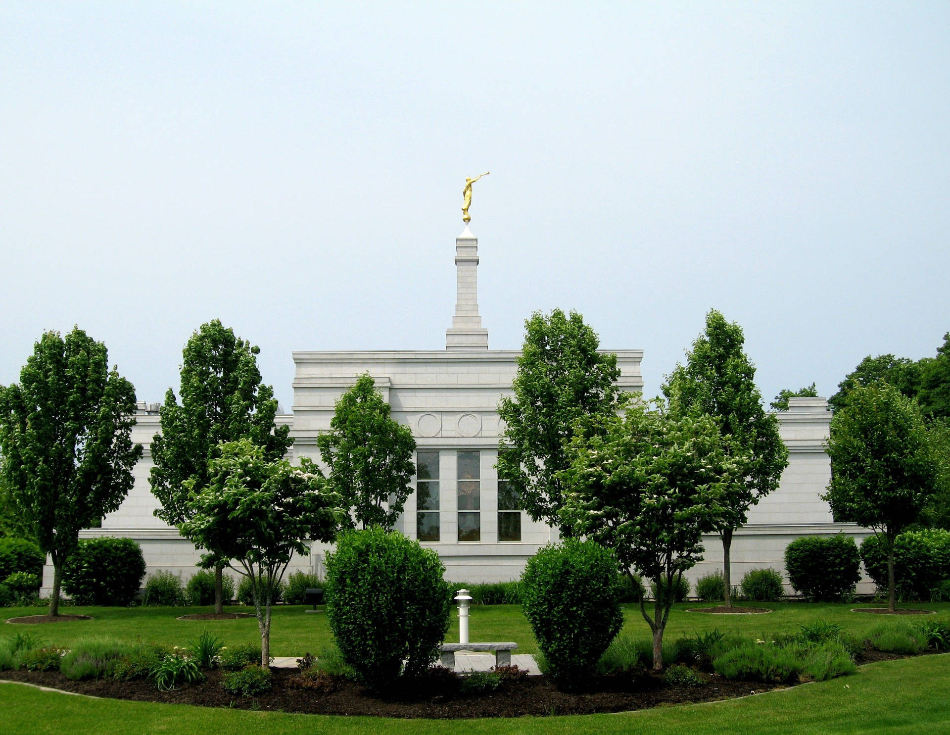 帕爾米拉紐約聖殿正面 壁紙 — 一座宏偉的白色寺廟，其尖頂上有一座金色雕像，在陰天之下，寧靜地矗立在茂密的綠樹和修剪整齊的草坪之中。
