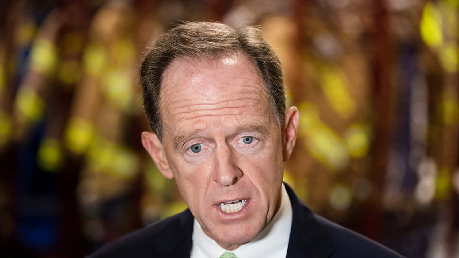 帕特·圖米低頭望向記者 壁紙 — 身穿西裝、擁有銳利藍眼的男士特寫肖像，背景為模糊的金色。