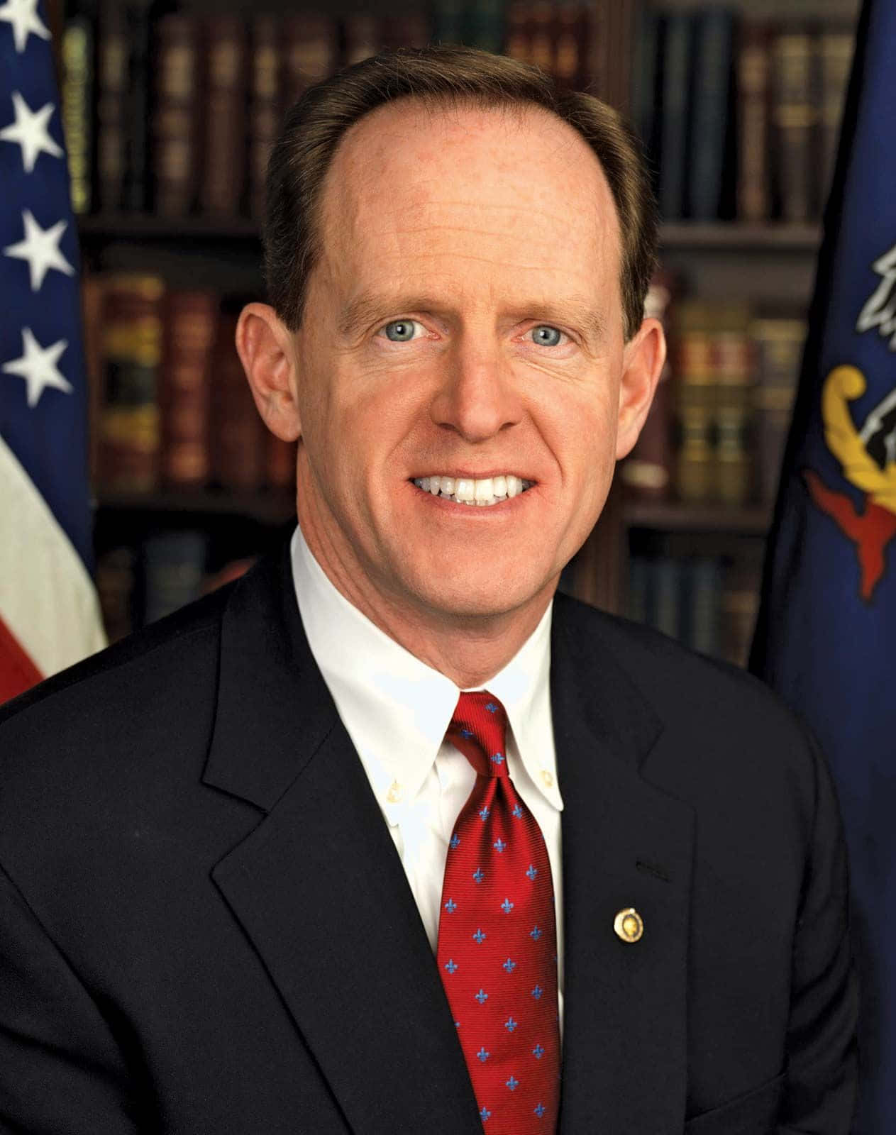 帕特·圖米參議院官方肖像 壁紙 — 一位微笑的男子身穿深色西裝和紅色圖案領帶，站在擺有美國國旗和州旗的書架前。