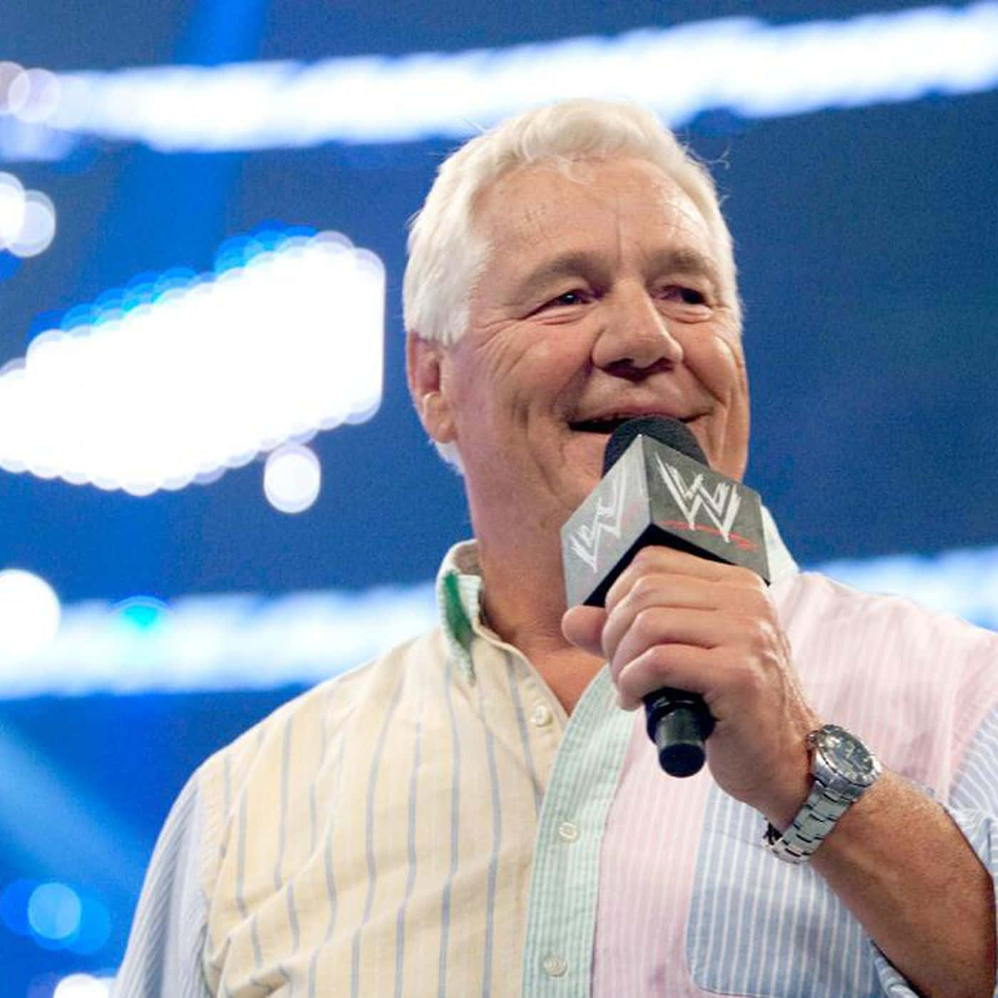 帕特·帕特森於 WWE 舞台發表介紹講話 壁紙 — 一位白髮老人在明亮的藍色舞台燈光下微笑著手持WWE麥克風。