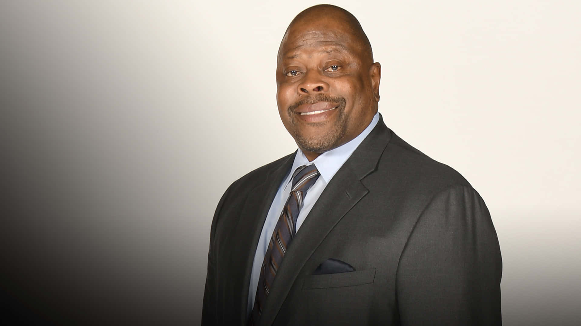 帕特里克·尤因NBA名人堂球員 壁紙 — 一位微笑的光頭黑人男性，身穿深色西裝和條紋領帶，在柔和的漸變工作室背景前自信地擺姿勢。