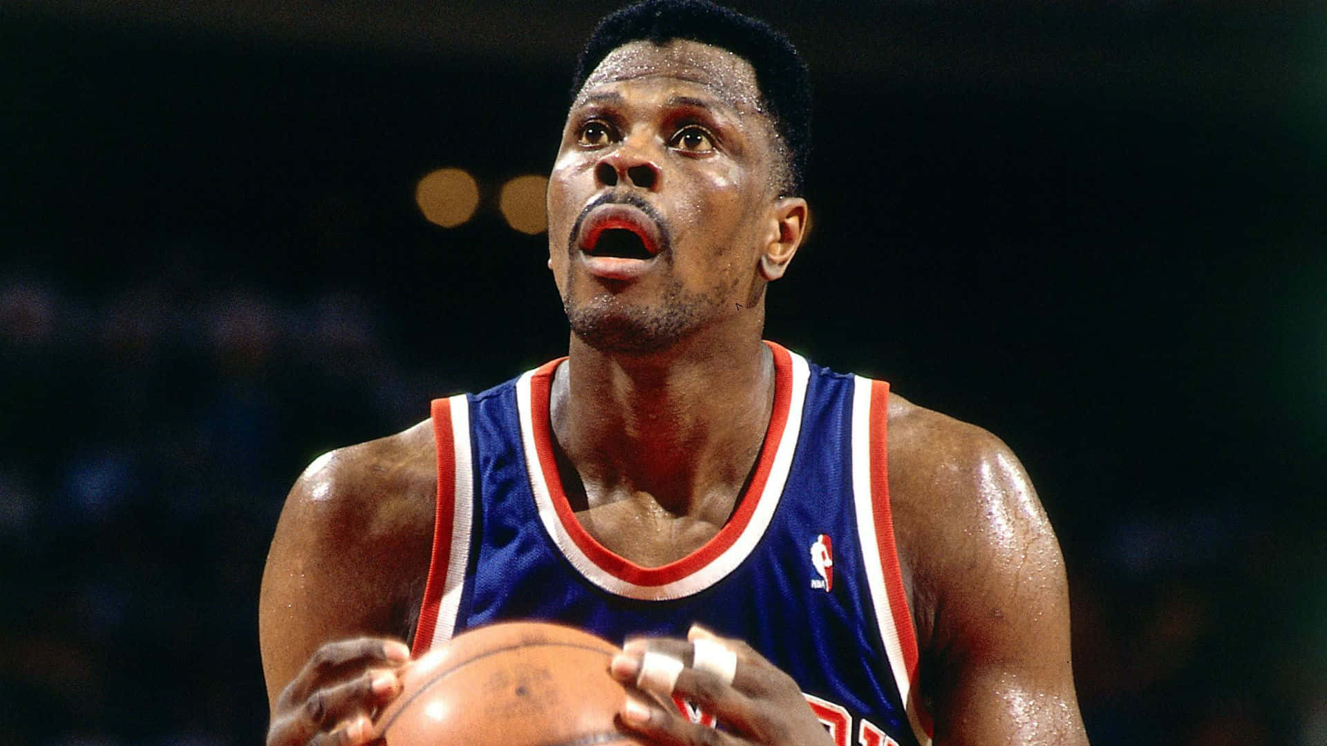 帕特里克·尤因NBA籃球中鋒 壁紙 — 一位專注的籃球運動員身穿藍色球衣，在場館燈光下於比賽中抓住球。
