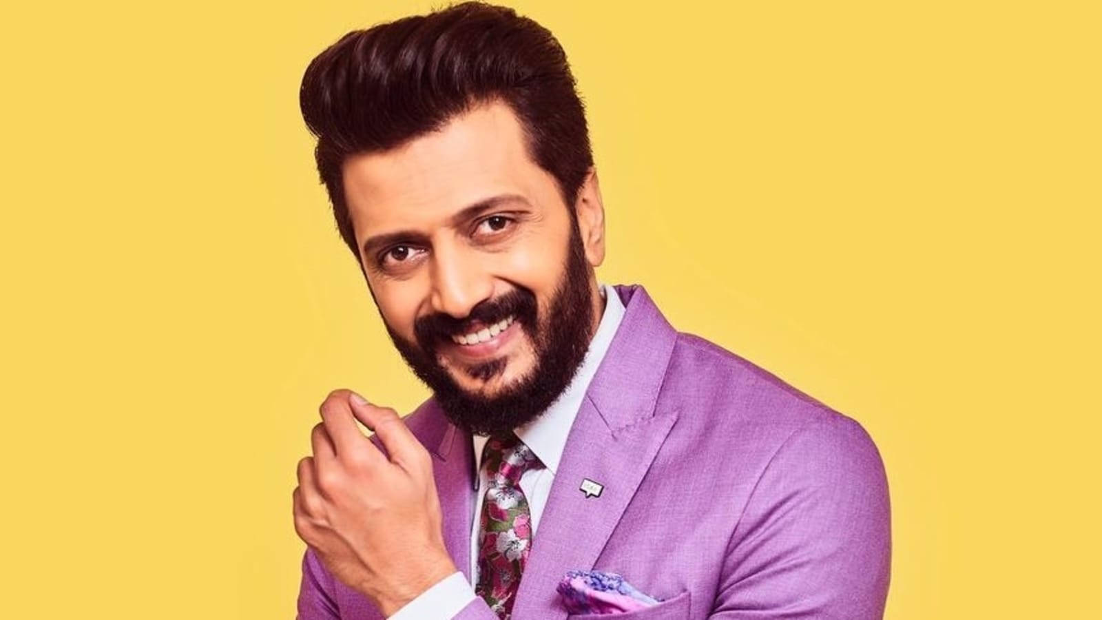 帥氣 Riteish Deshmukh 身穿粉紅西裝 壁紙 — 一位留鬍鬚的微笑男士穿著紫色西裝，背景是明亮的黃色。