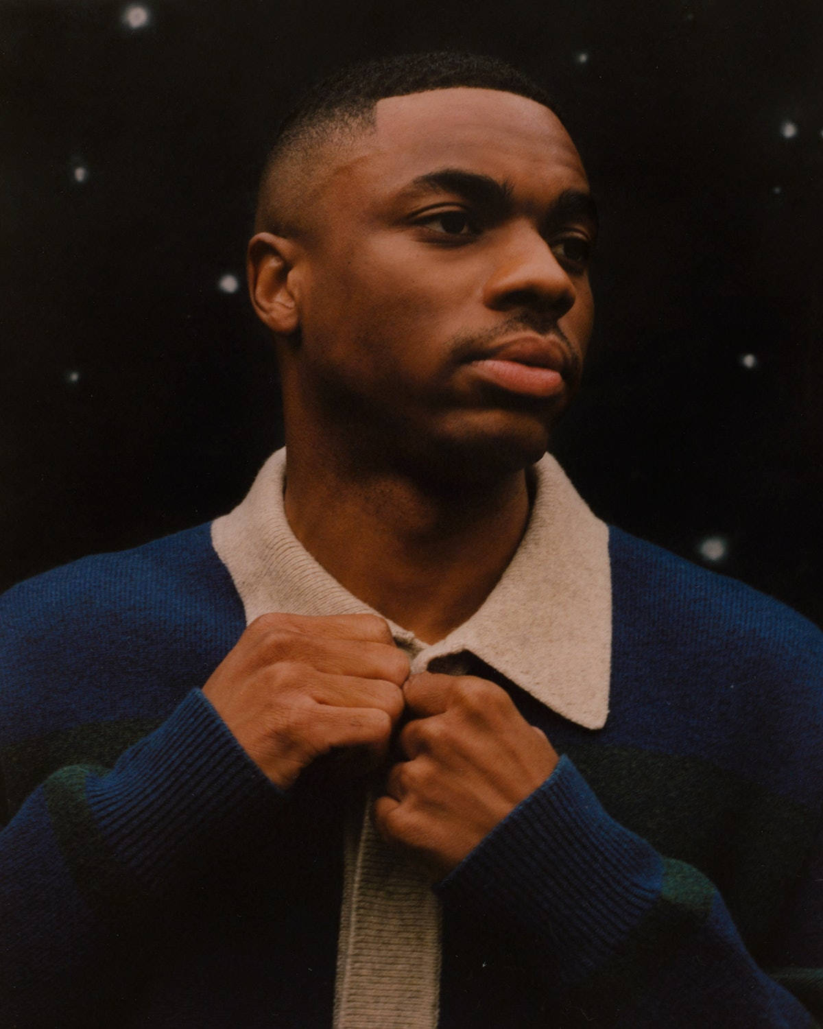 帥氣 Vince Staples 壁紙 — 一位身穿藍色和米色針織毛衣的年輕男子在星空夜空背景下調整衣領。