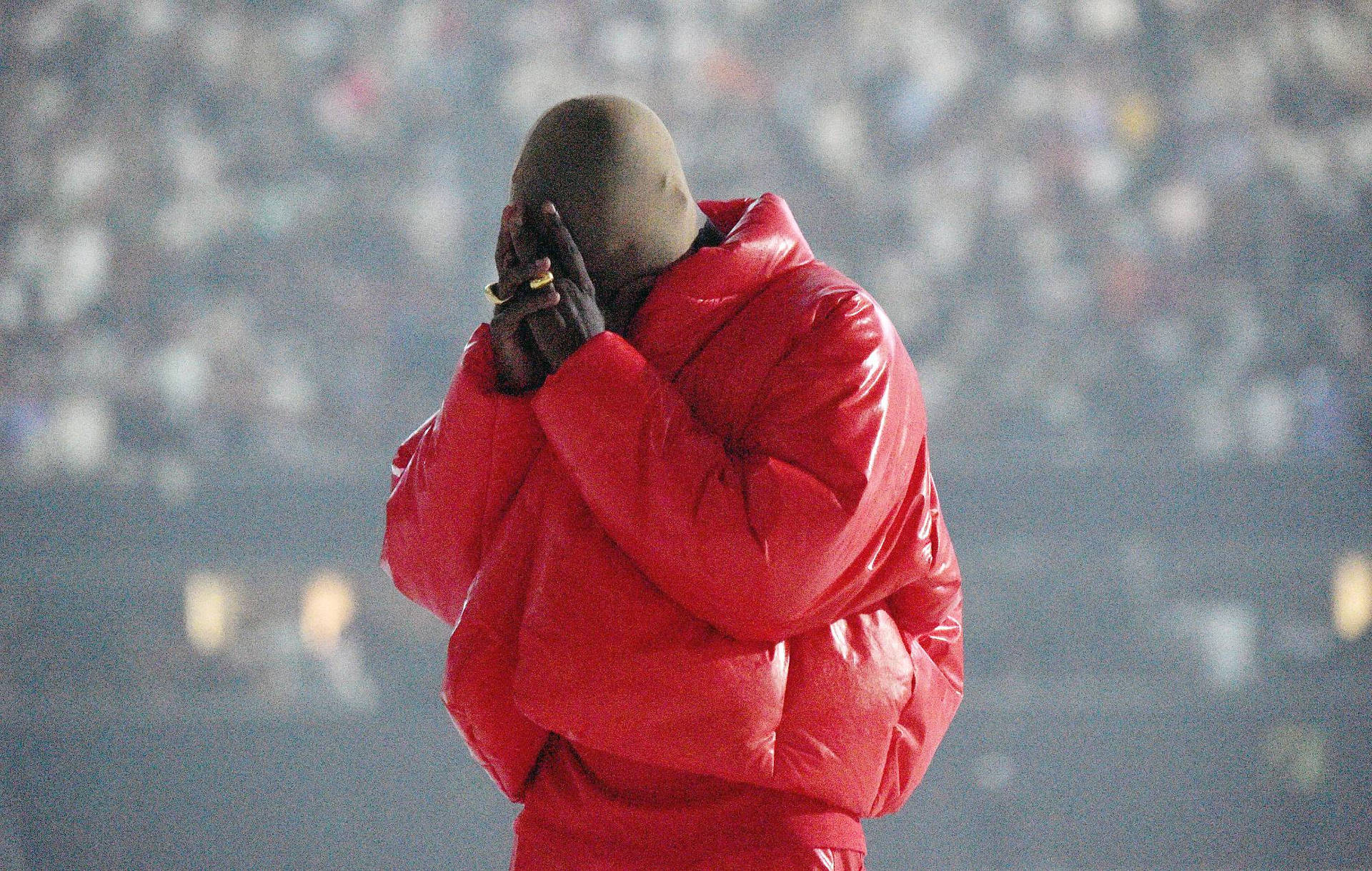 帥氣坎耶·威斯特 Donda 紅色 Yeezy 壁紙 — 一個穿著閃亮紅色羽絨外套的男人背對鏡頭站立，雙手遮臉，背景是模糊的人群。