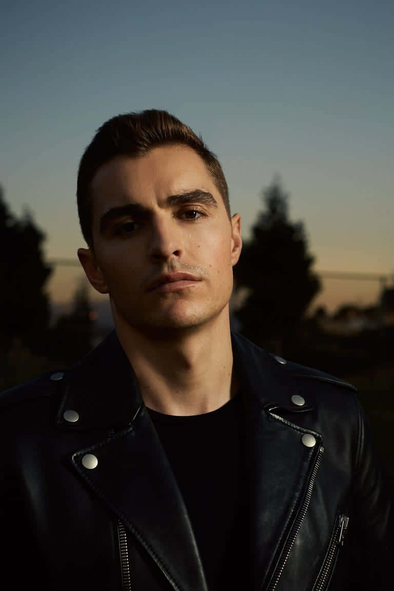 帥氣男士 Dave Franco 壁紙 — 一個穿著黑色皮褸的男人，在模糊的樹木背景下，對著柔和嘅暮色天空專注地凝視。