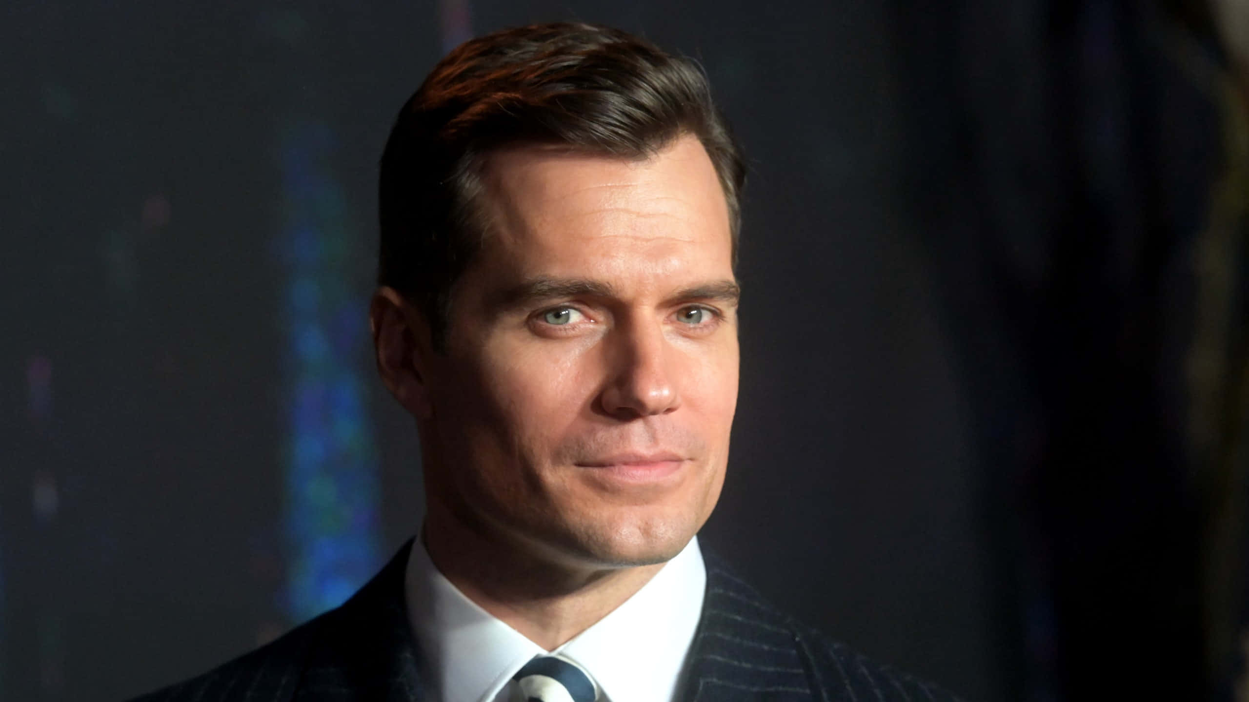 帥氣男士 Henry Cavill 嘲笑表情 壁紙 — 一個穿著細條紋西裝的男人在黑暗模糊背景下的特寫肖像，帶著微妙微笑。